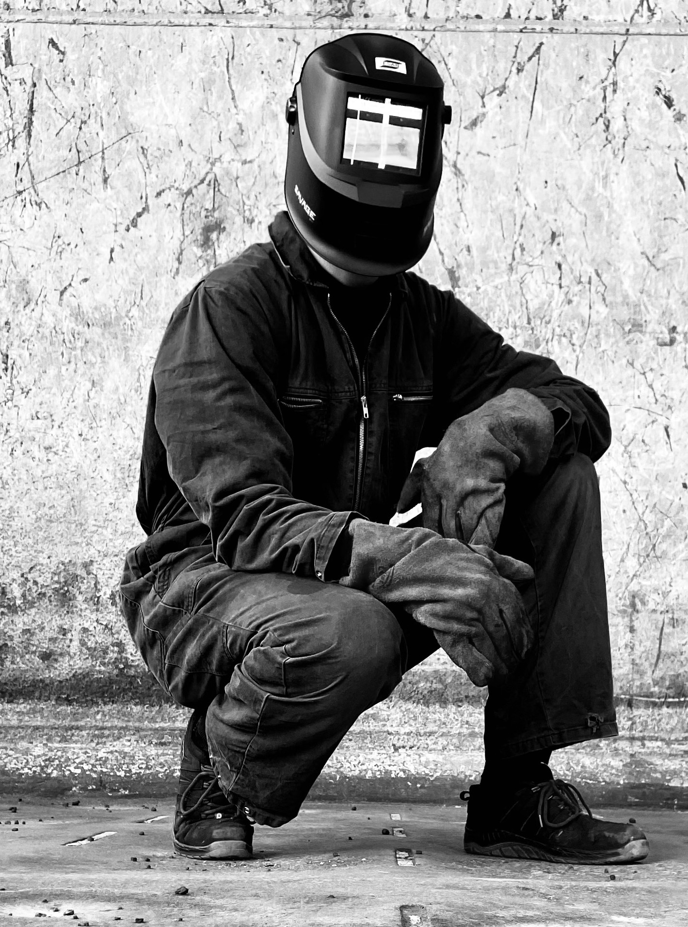 Czarno-białe zdjęcie spawacza w kombinezonie i masce ochronnej, w pozycji kucznej, z rękawicami roboczymi na kolanach. Tło stanowi surowa, teksturowana ściana.