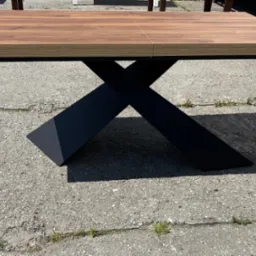 Rozkładany stół z drewnianym blatem i czarnymi, metalowymi nogami w kształcie litery X, stojący na betonowej nawierzchni. Widoczne detale łączenia blatu.