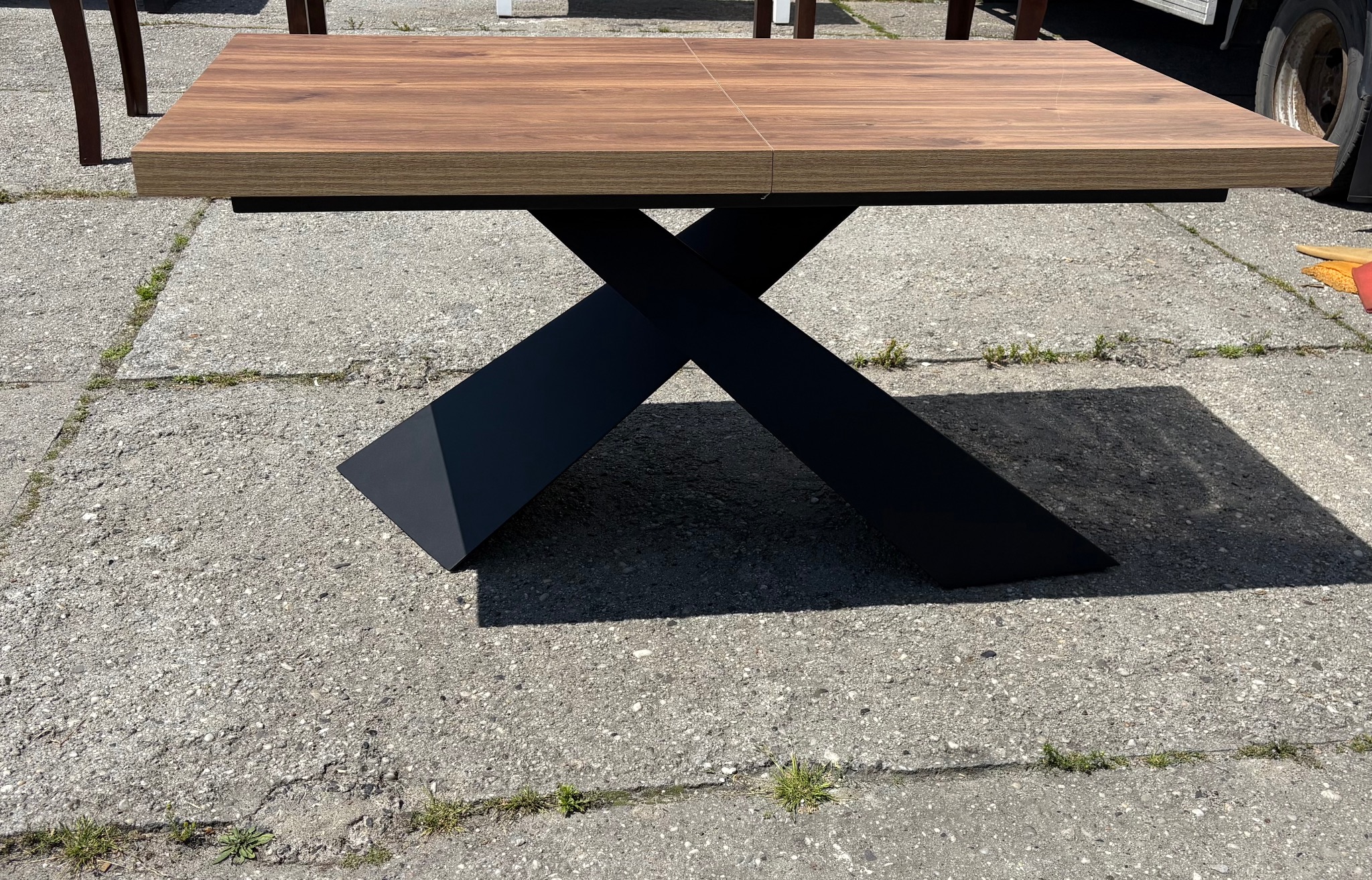 Rozkładany stół z drewnianym blatem i czarnymi, metalowymi nogami w kształcie litery X, stojący na betonowej nawierzchni. Widoczne detale łączenia blatu.
