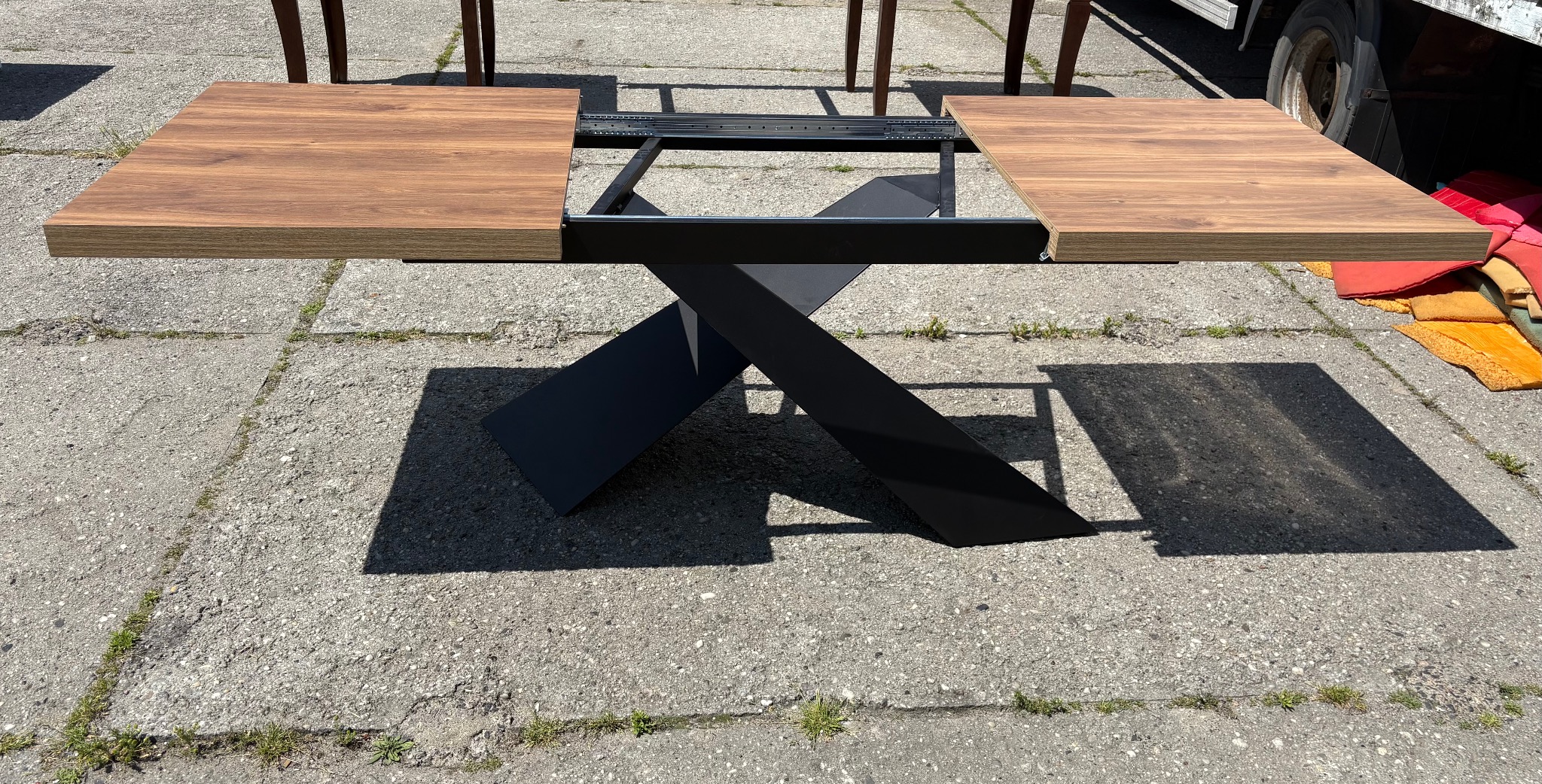 Rozkładany stół z drewnianym blatem i czarną, metalową podstawą w kształcie litery X, ustawiony na betonowej nawierzchni. Widoczne mechanizmy rozkładania blatu.