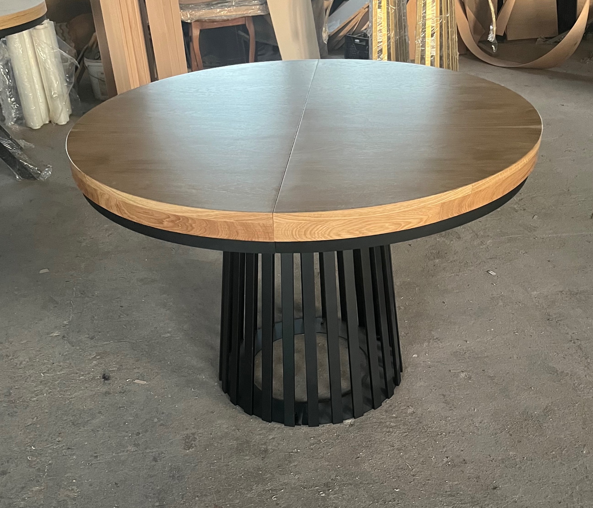 Okrągły stół z drewnianym blatem i czarną, metalową podstawą w kształcie klatki. Blat składa się z dwóch części, widoczne łączenie na środku.