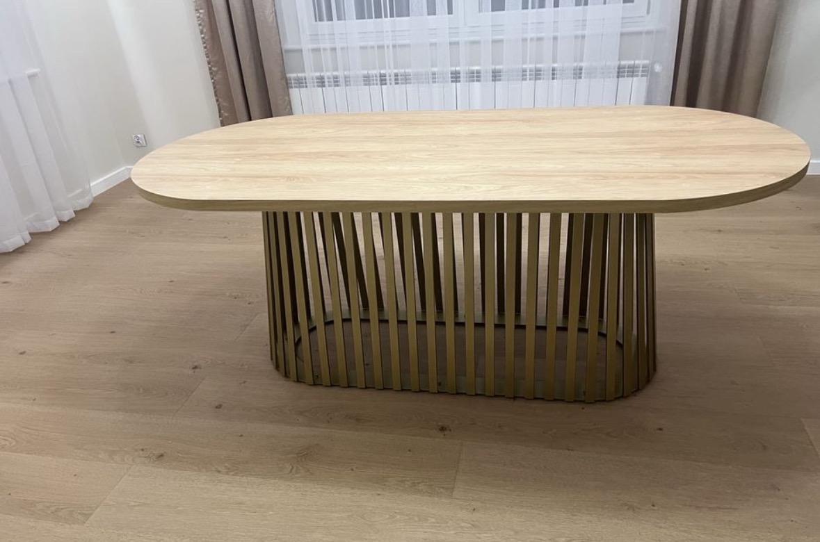 Stół z blatem z jasnego drewna i metalową podstawą w kształcie ażurowej klatki. Nowoczesny design, widok z boku, na drewnianej podłodze, zasłony w tle.