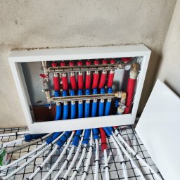 Tomasz Gorlowski - Rozdzielacz ogrzewania podłogowego w białej obudowie, z rurami w izolacji czerwonej i niebieskiej, ułożonymi na folii izolacyjnej z siatką. Surowy stan ścian.