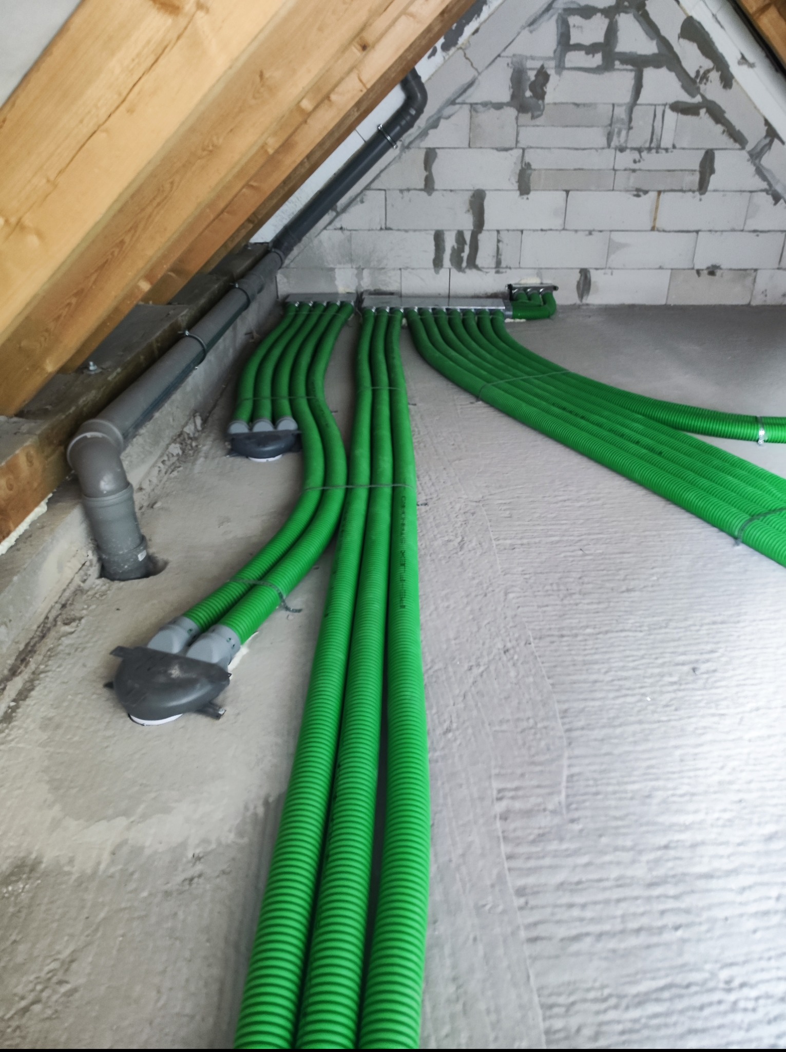 Zielone rury wentylacyjne ułożone na poddaszu w budynku z drewnianą konstrukcją dachu i ścianami z betonu komórkowego, widoczne elementy instalacji kanalizacyjnej.