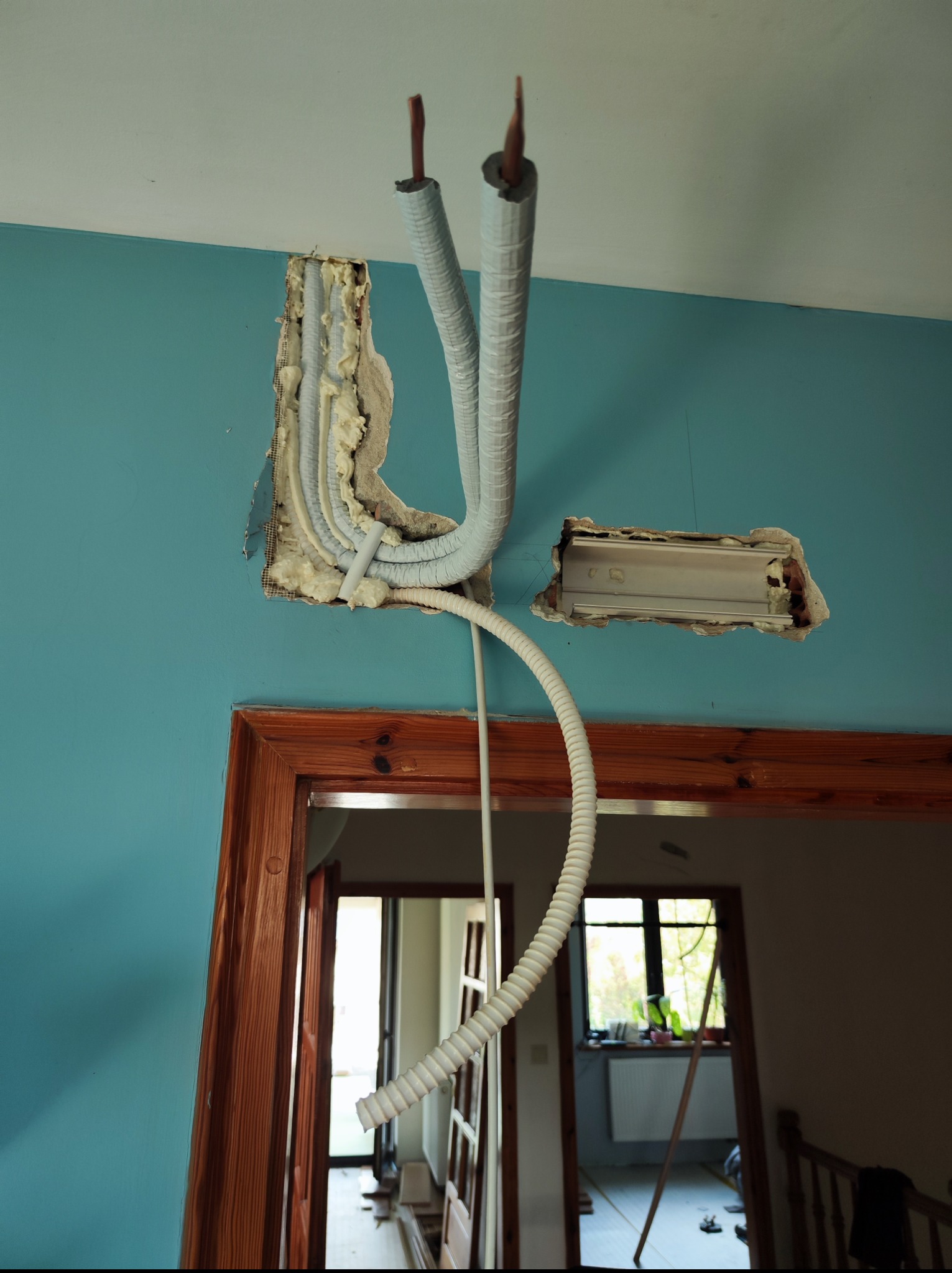 Instalacja przewodów w ścianie o turkusowym odcieniu, zabezpieczonych białą izolacją, wystających z otworów wypełnionych pianką montażową. Widok na wnętrze przez drewniane futryny.