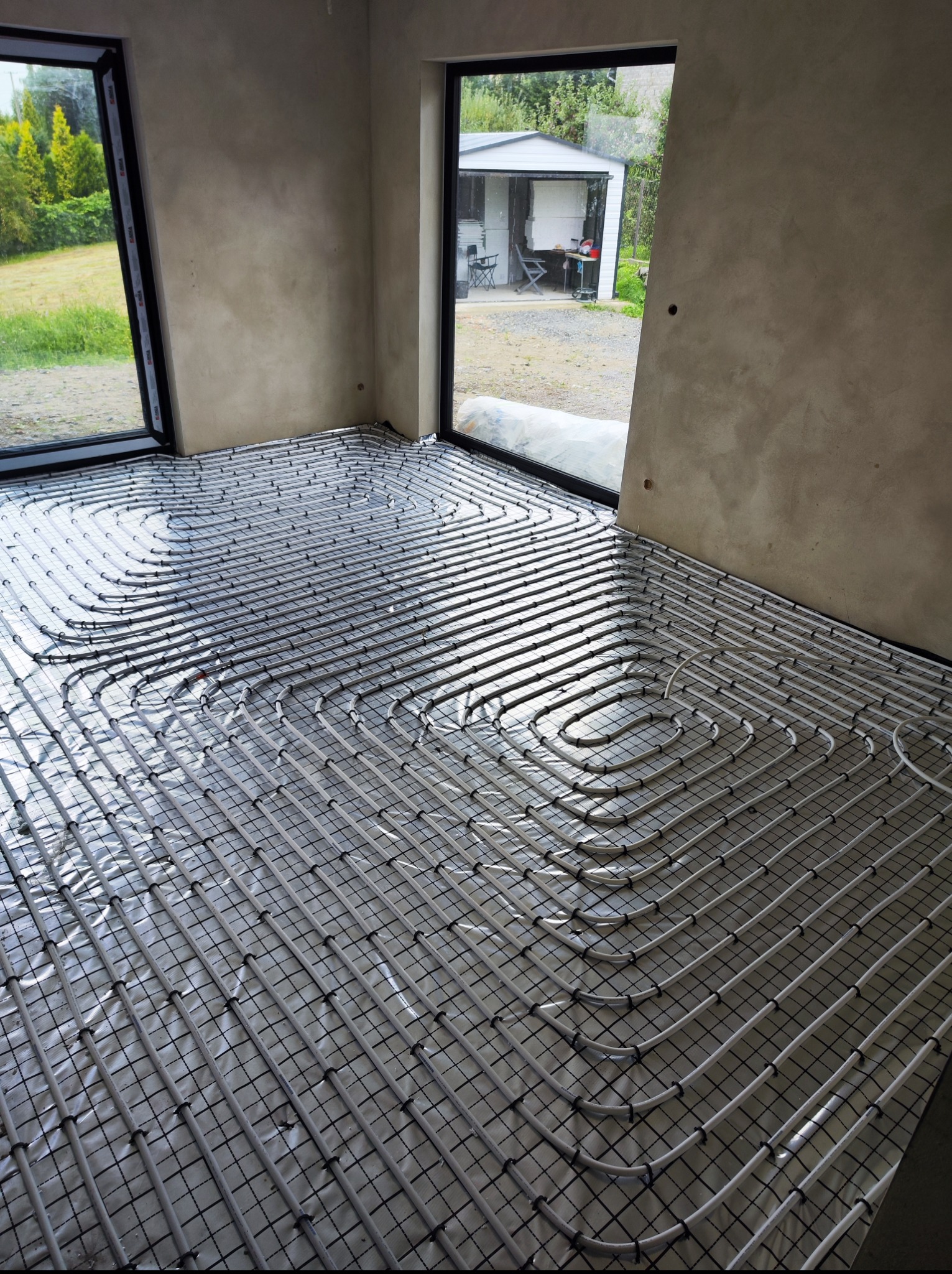 Białe rury ogrzewania podłogowego ułożone na folii, widok z góry, surowe ściany i okna z widokiem na zewnątrz. Instalacja w trakcie realizacji.