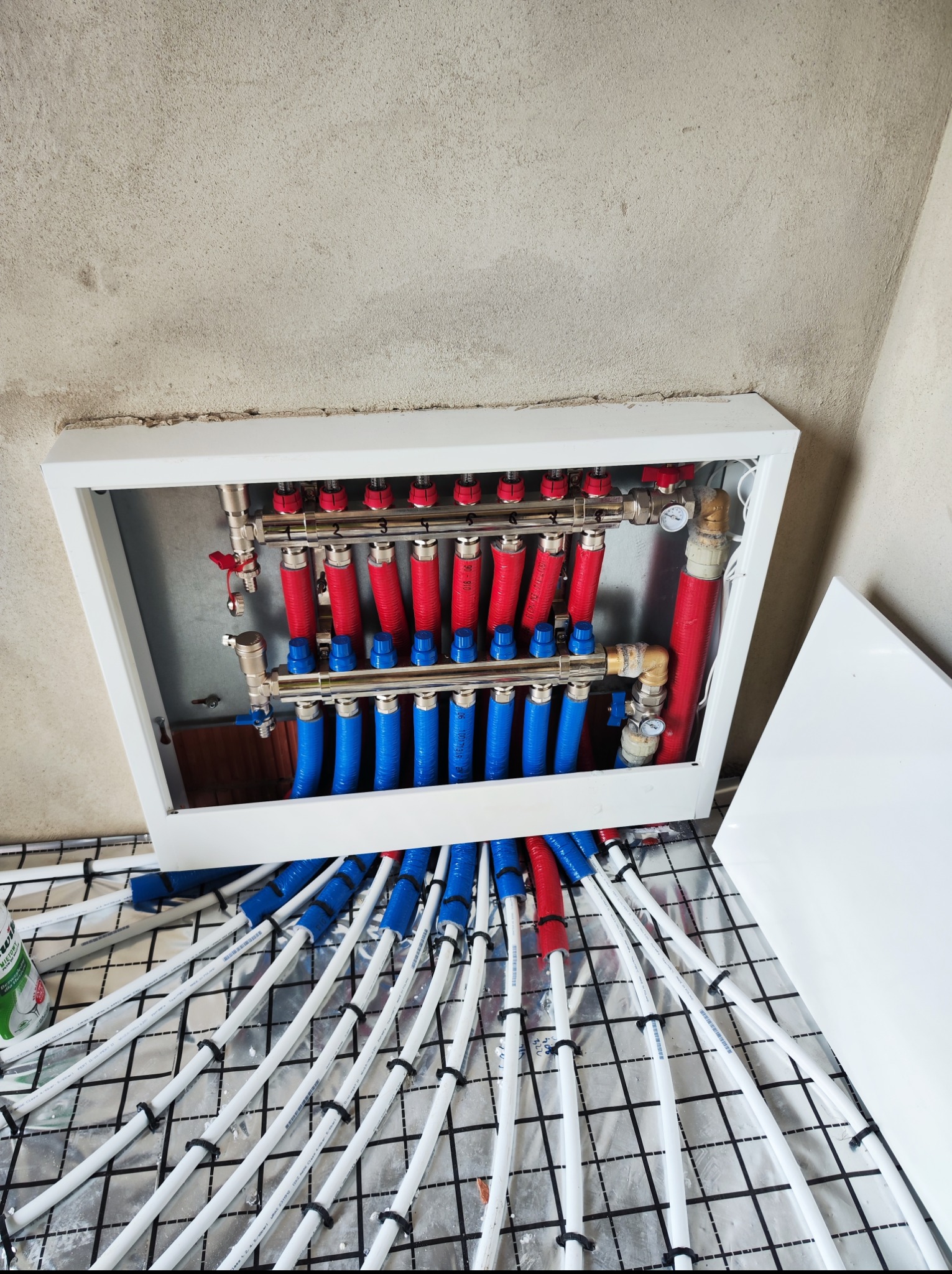 Rozdzielacz ogrzewania podłogowego w białej obudowie, z rurami w izolacji czerwonej i niebieskiej, ułożonymi na folii izolacyjnej z siatką. Surowy stan ścian.