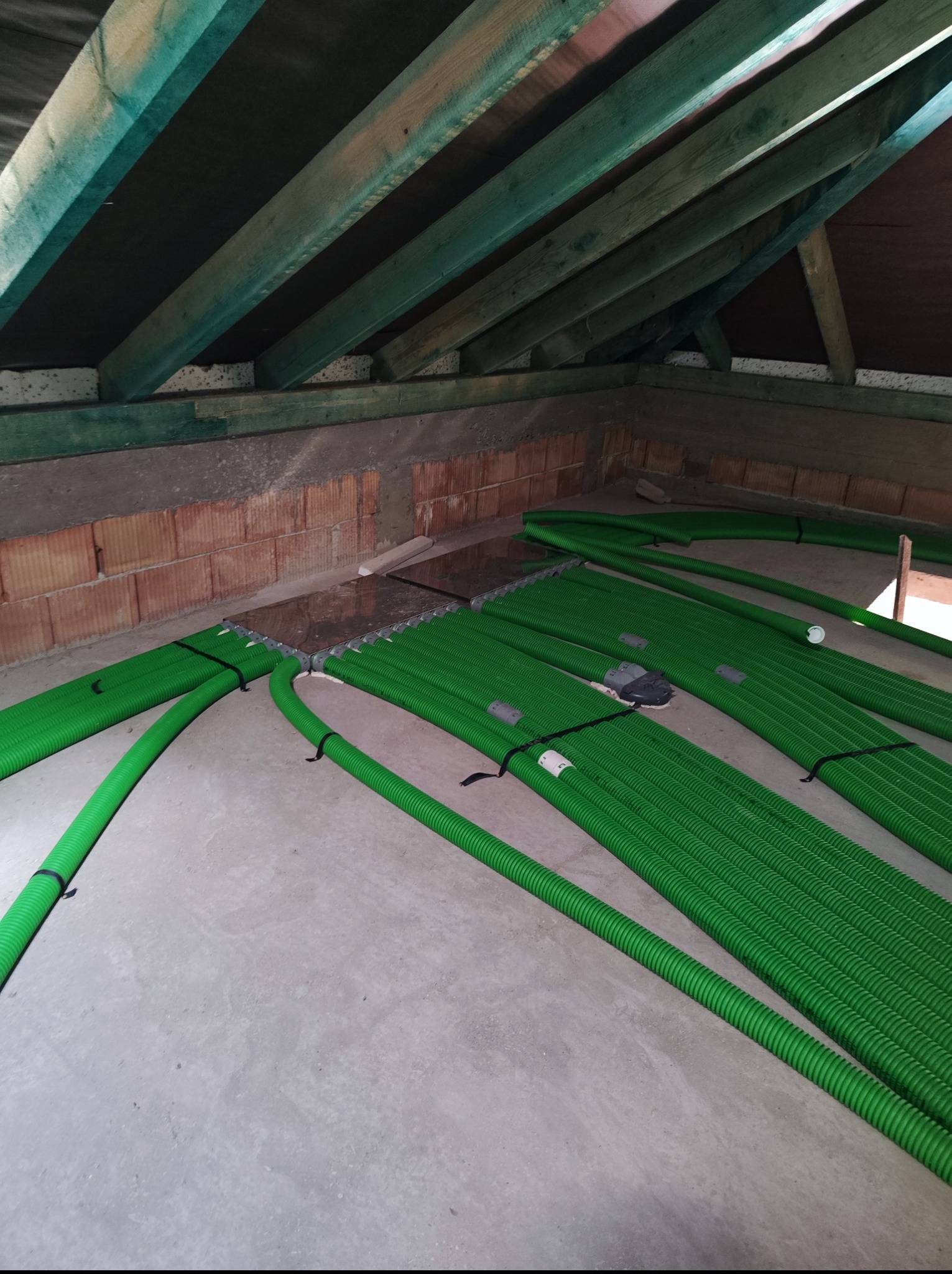 Zielone rury ogrzewania podłogowego ułożone na szarej posadzce w surowym wnętrzu poddasza, widoczne drewniane krokwie i fragment muru z cegieł.