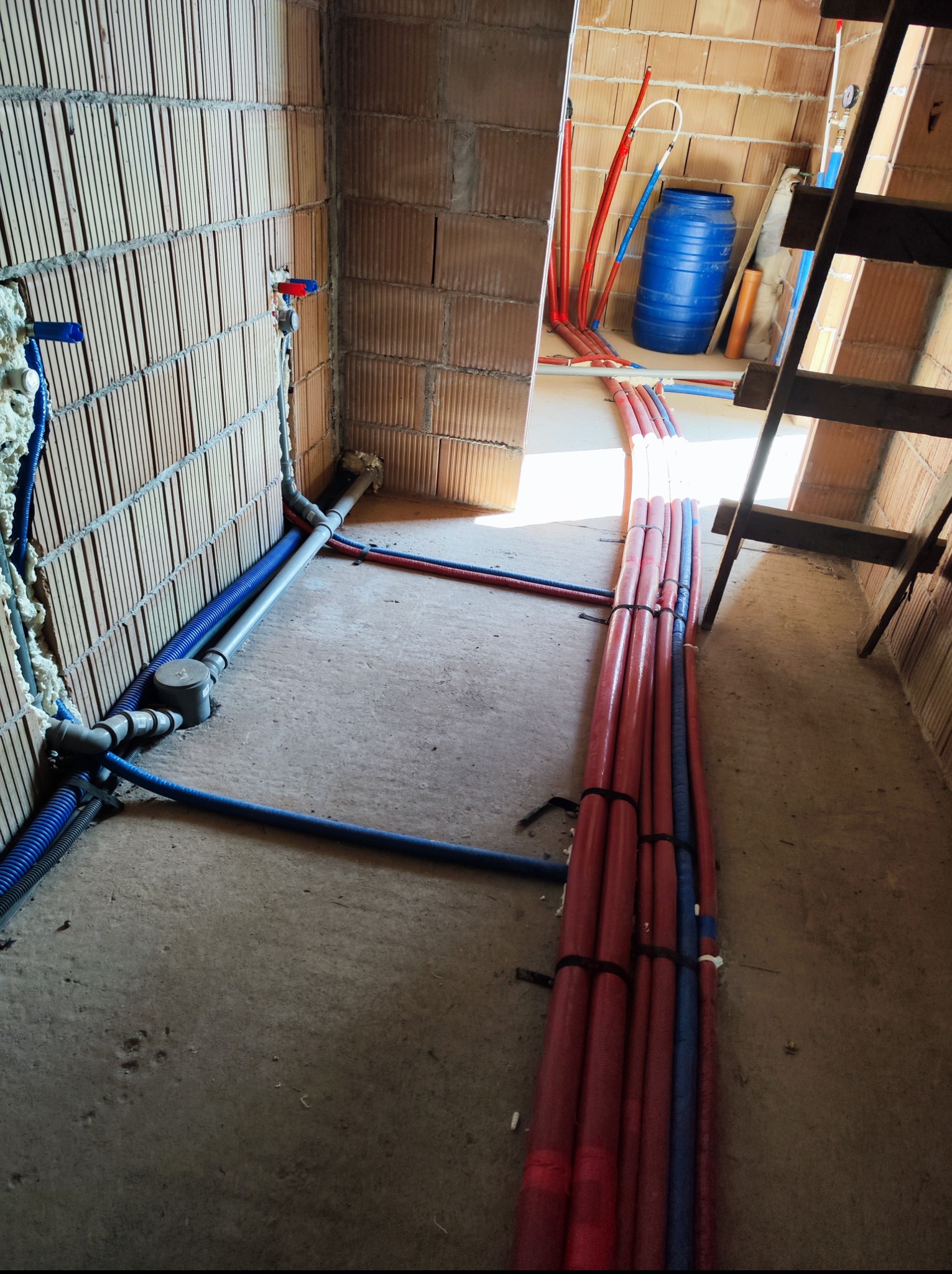 Surowy stan: instalacja wodna i grzewcza w budynku z cegły. Rury czerwone i niebieskie prowadzone po podłodze, część w ścianie. Widoczna drabina i niebieska beczka.