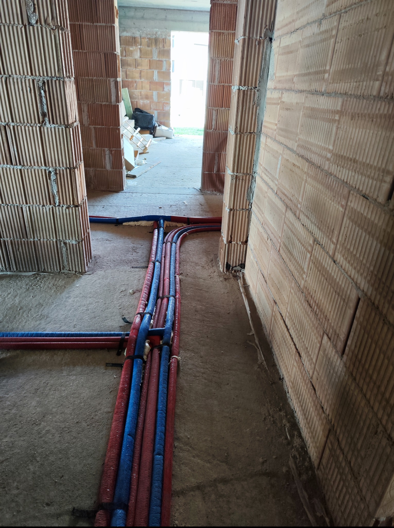Instalacja rur w budynku w trakcie budowy. Rury w izolacji czerwonej i niebieskiej ułożone wzdłuż ściany z cegły dziurawki. Widok na surowy stan budynku.