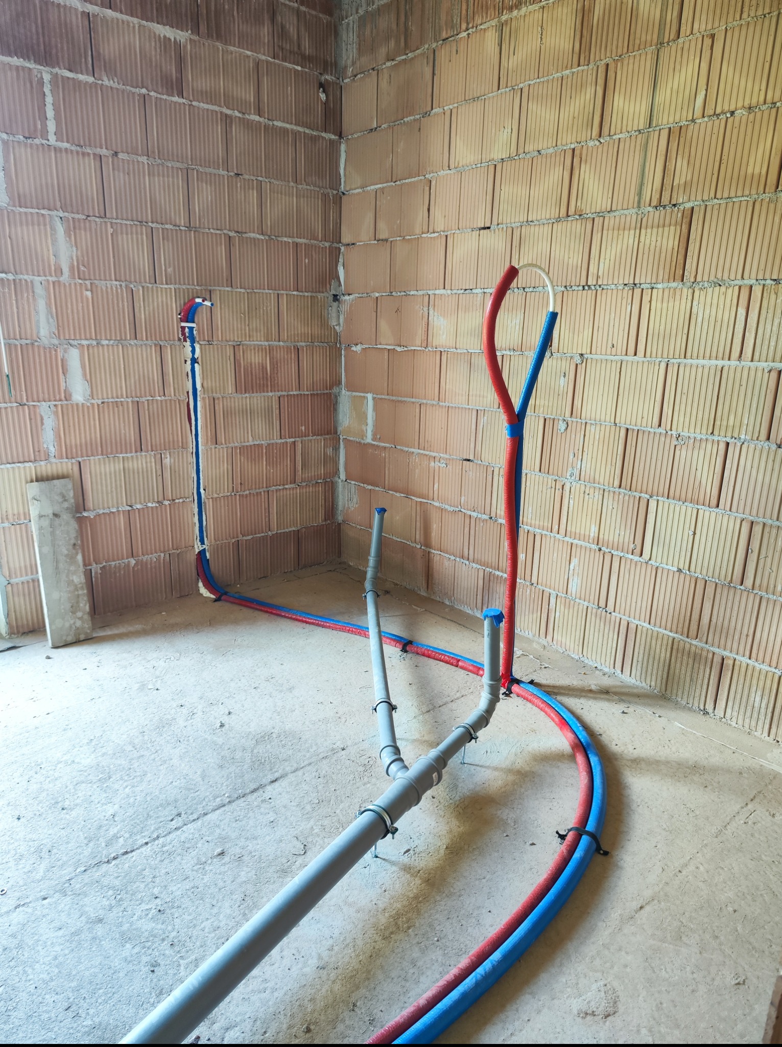Surowy mur z instalacją wodną i kanalizacyjną, rury w kolorach niebieskim i czerwonym. Widoczne łączenia i mocowania. Stan surowy, przed wykończeniem.