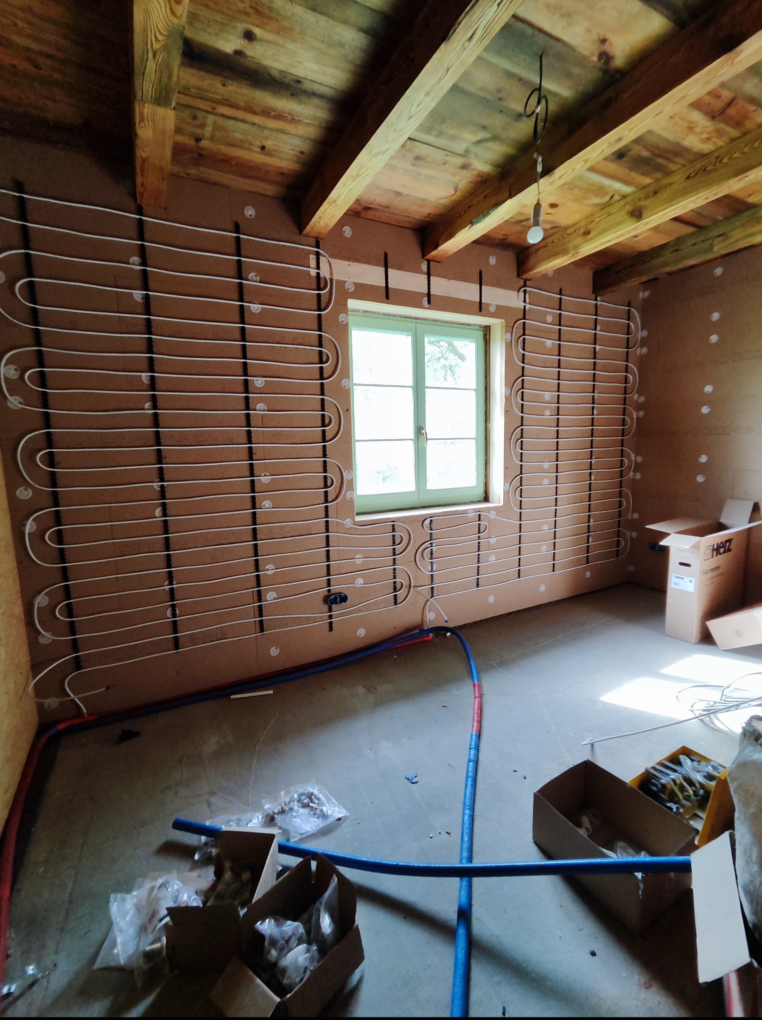 Instalacja ogrzewania podłogowego na ścianach w pomieszczeniu z drewnianym stropem. Widoczne rury i elementy montażowe, okno w tle.
