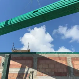 Ciężki dźwig podnosi pakiet impregnowanej, zielonej tarcicy budowlanej na tle błękitnego nieba i surowej konstrukcji budynku z czerwonej cegły.