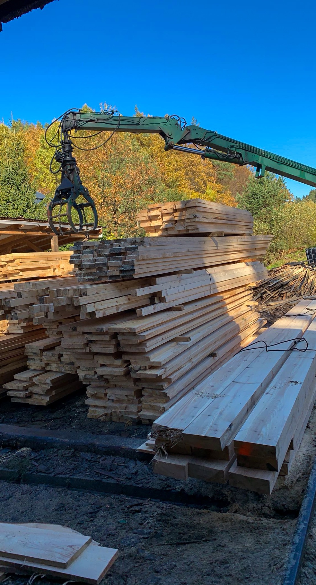 Stos tarcicy budowlanej ładowany przez dźwig z chwytakiem na tle jesiennego lasu i błękitnego nieba. Widoczne świeże drewno i fragmenty innych stosów.