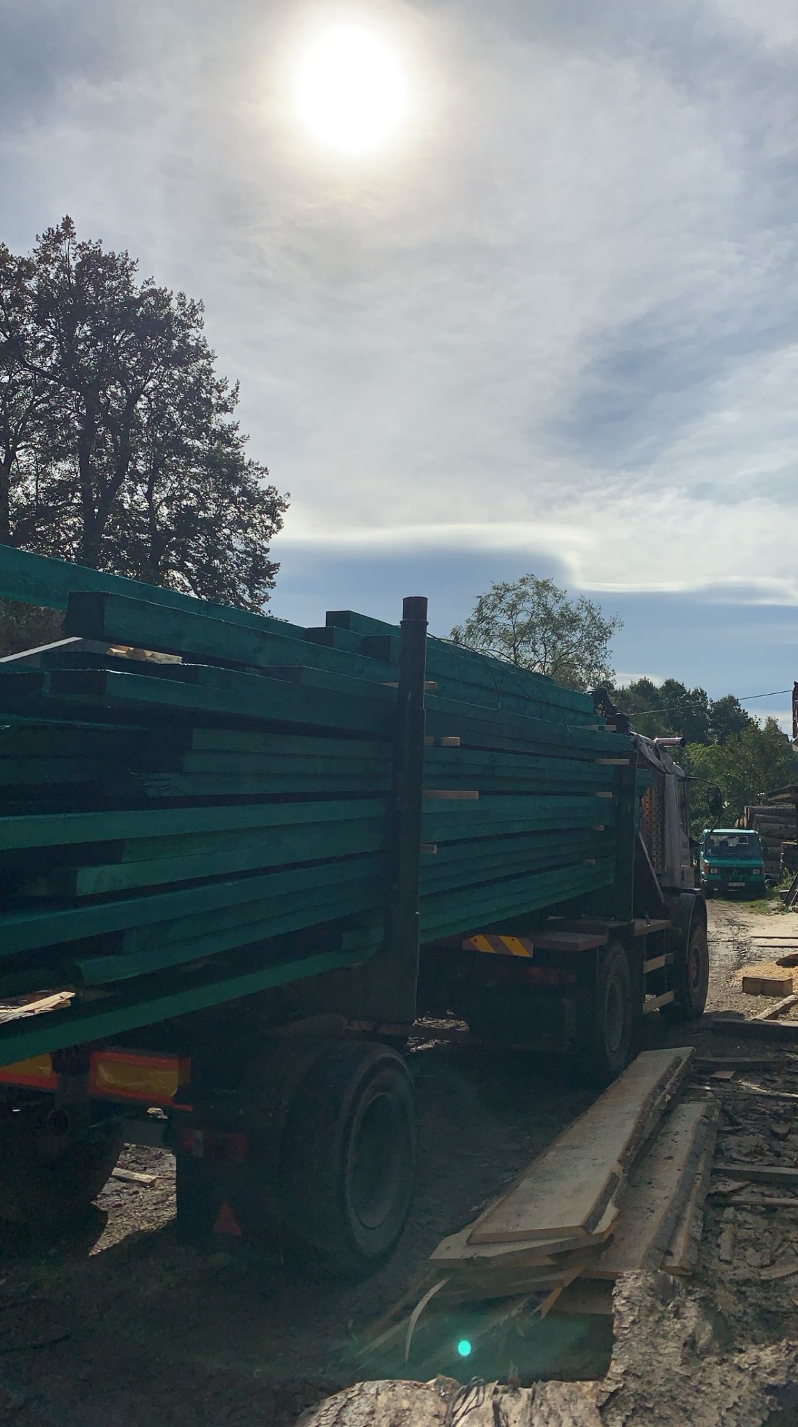 Ciężarówka z ładunkiem zielonej tarcicy na tle drzew i nieba. Na pierwszym planie ułożone deski.