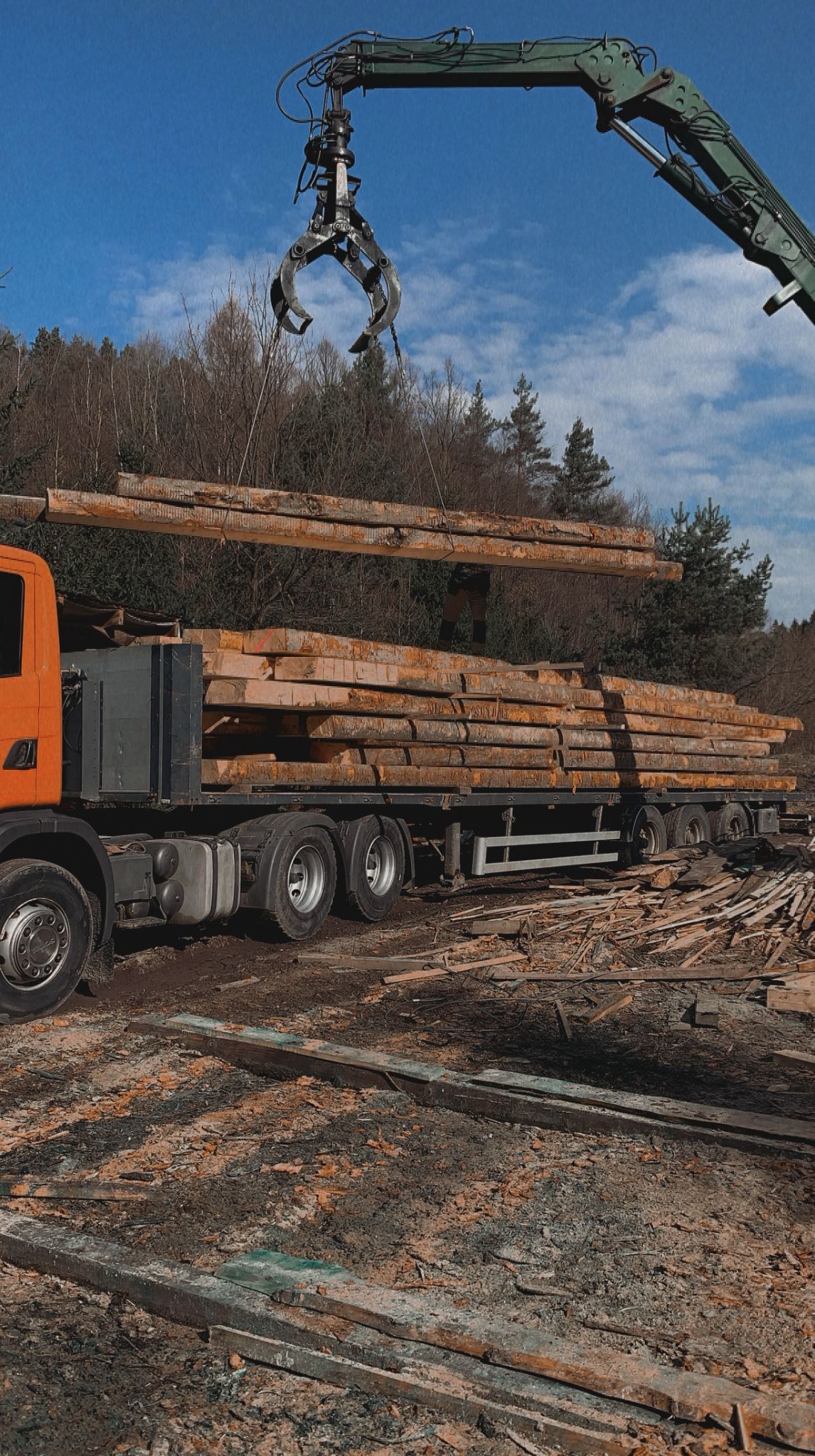 Załadunek tarcicy na ciężarówkę za pomocą chwytaka. Widoczne drewniane bale, ciężarówka z przyczepą i chwytak na tle lasu i błękitnego nieba.