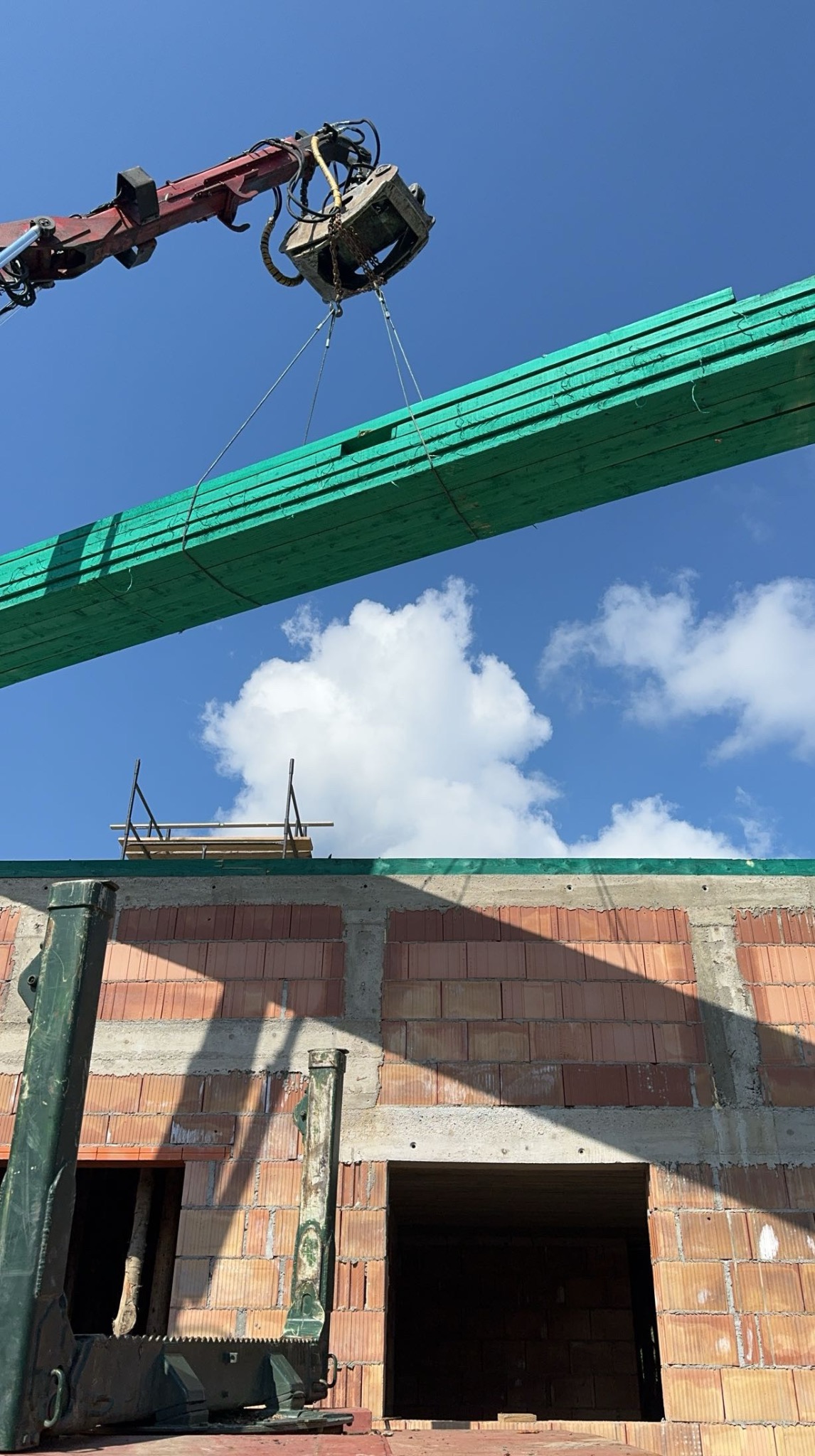 Ciężki dźwig podnosi pakiet impregnowanej, zielonej tarcicy budowlanej na tle błękitnego nieba i surowej konstrukcji budynku z czerwonej cegły.