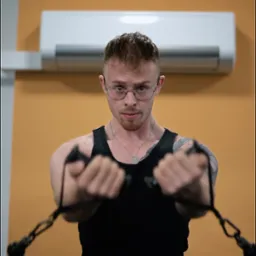 Mężczyzna w okularach ćwiczy na maszynie z linkami w siłowni; widoczny drążek do podciągania i klimatyzator. Koncentracja na ćwiczeniu, trening siłowy.