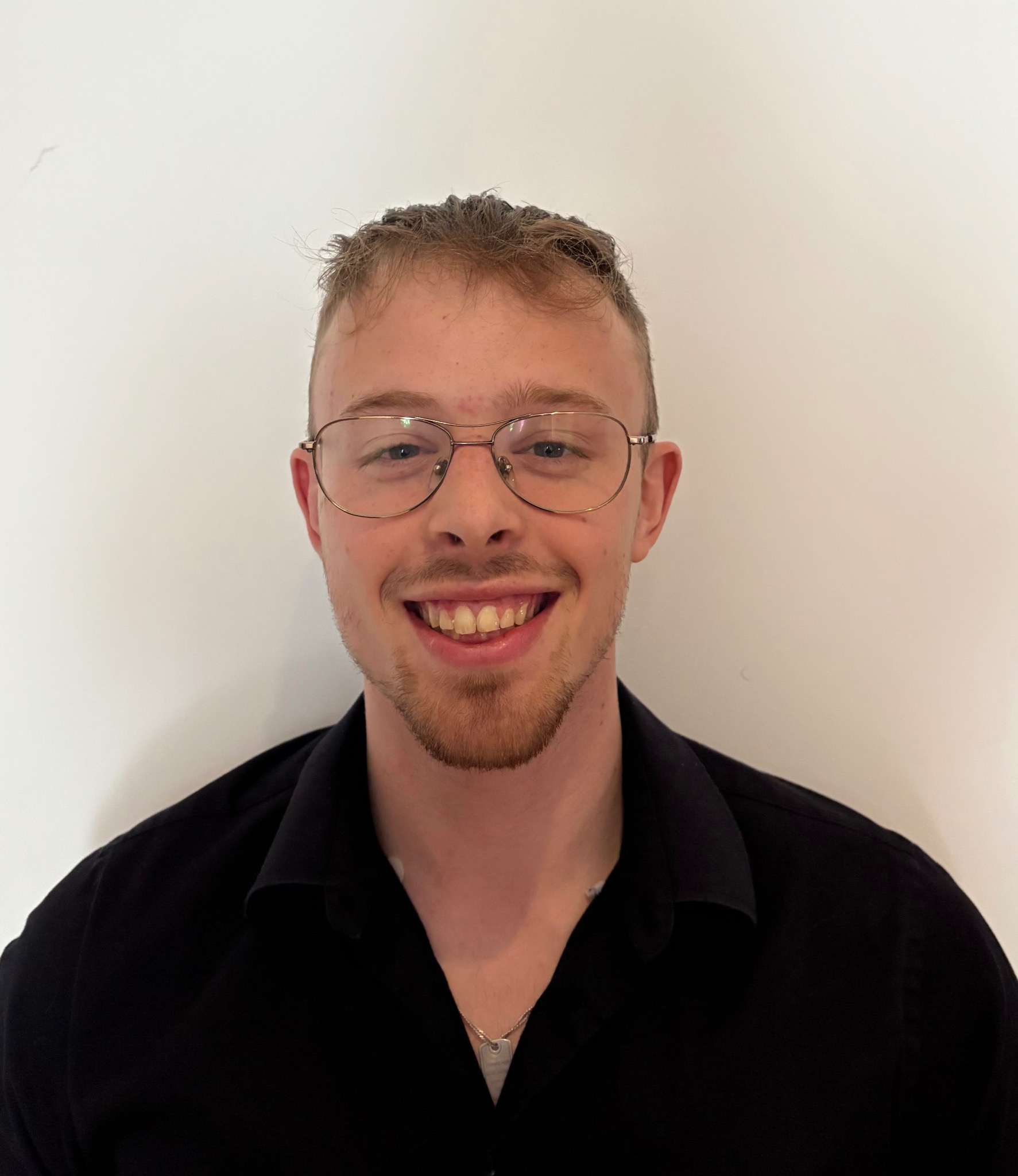 Portret uśmiechniętego mężczyzny w okularach i czarnej koszuli na tle białej ściany. Widoczne delikatne zmarszczki mimiczne i srebrny łańcuszek na szyi.