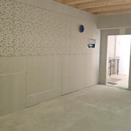 Jankowski - Wnętrze w trakcie montażu płyt gipsowo-kartonowych. Widoczne płyty o różnej fakturze, surowy beton na podłodze i drewniane belki stropowe. Otwór drzwiowy w tle.