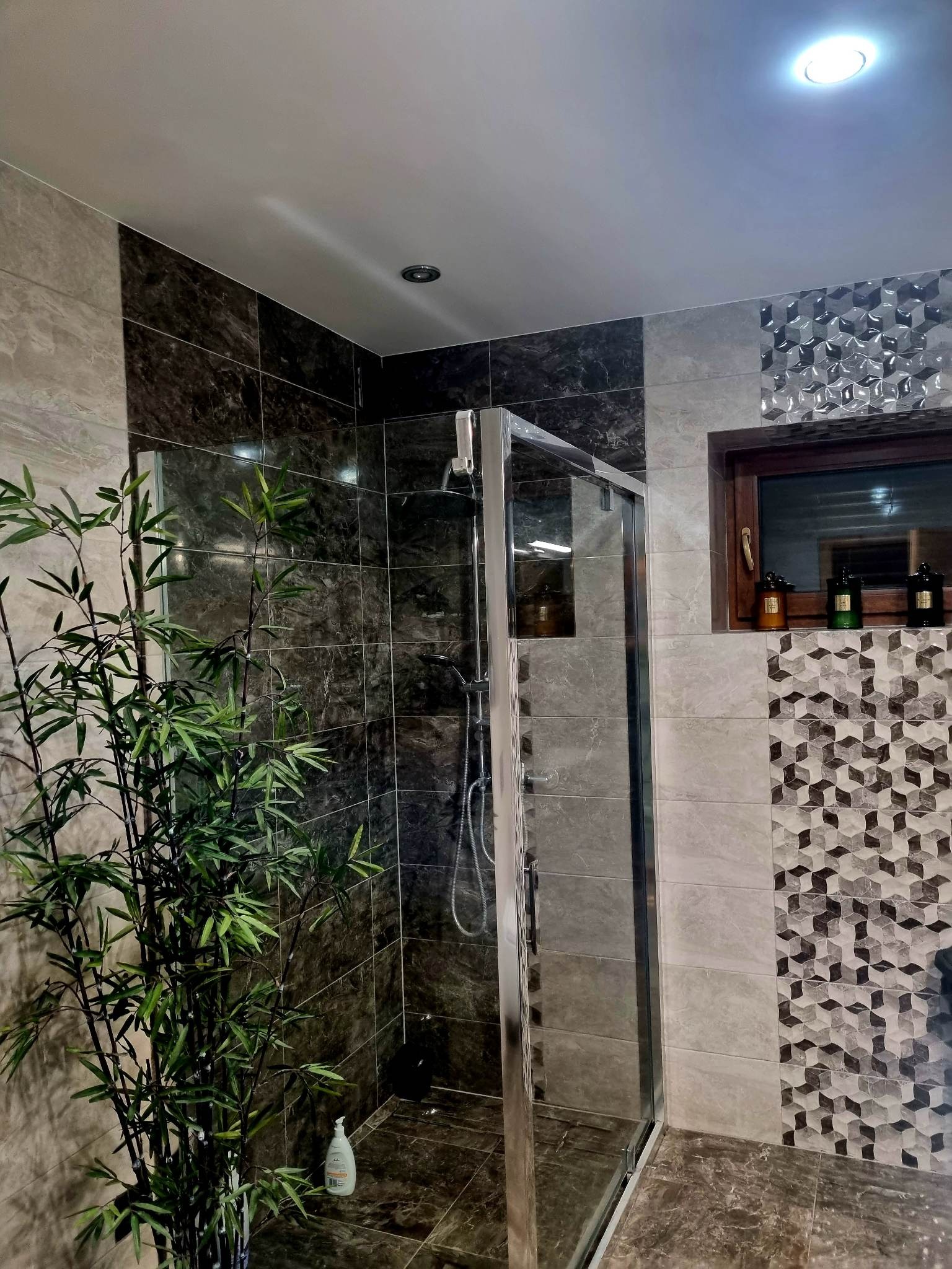 Nowoczesna łazienka z kabiną prysznicową, wykończona płytkami w odcieniach szarości i brązu. Dekoracyjne płytki geometryczne i bambus dodają charakteru.