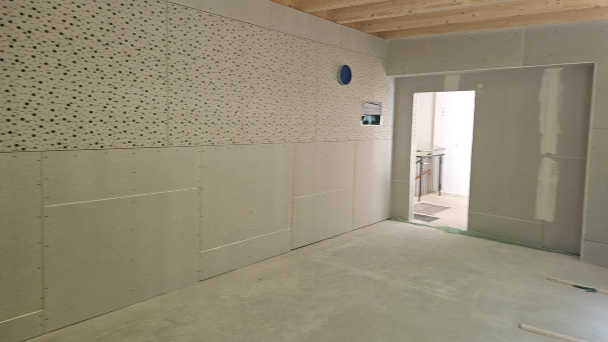 Wnętrze w trakcie montażu płyt gipsowo-kartonowych. Widoczne płyty o różnej fakturze, surowy beton na podłodze i drewniane belki stropowe. Otwór drzwiowy w tle.