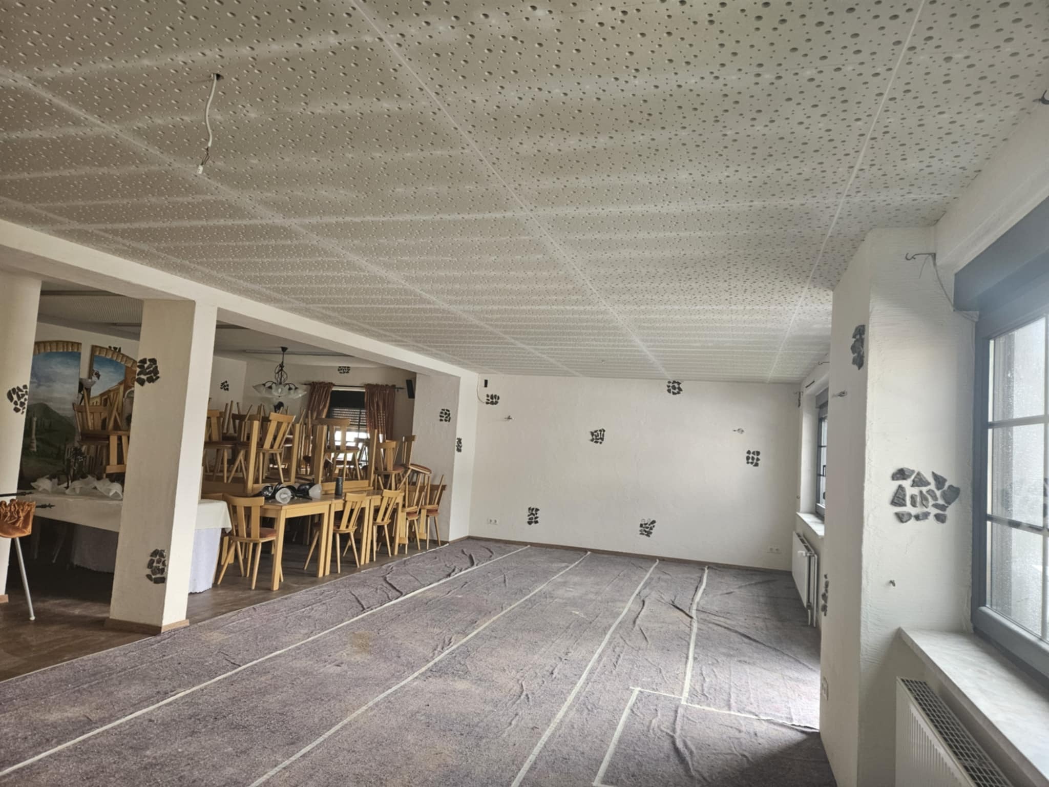 Przygotowanie sali do remontu: zabezpieczona podłoga, ściany z dekoracyjnymi elementami, perforowany sufit podwieszany i ustawione stoły z krzesłami.