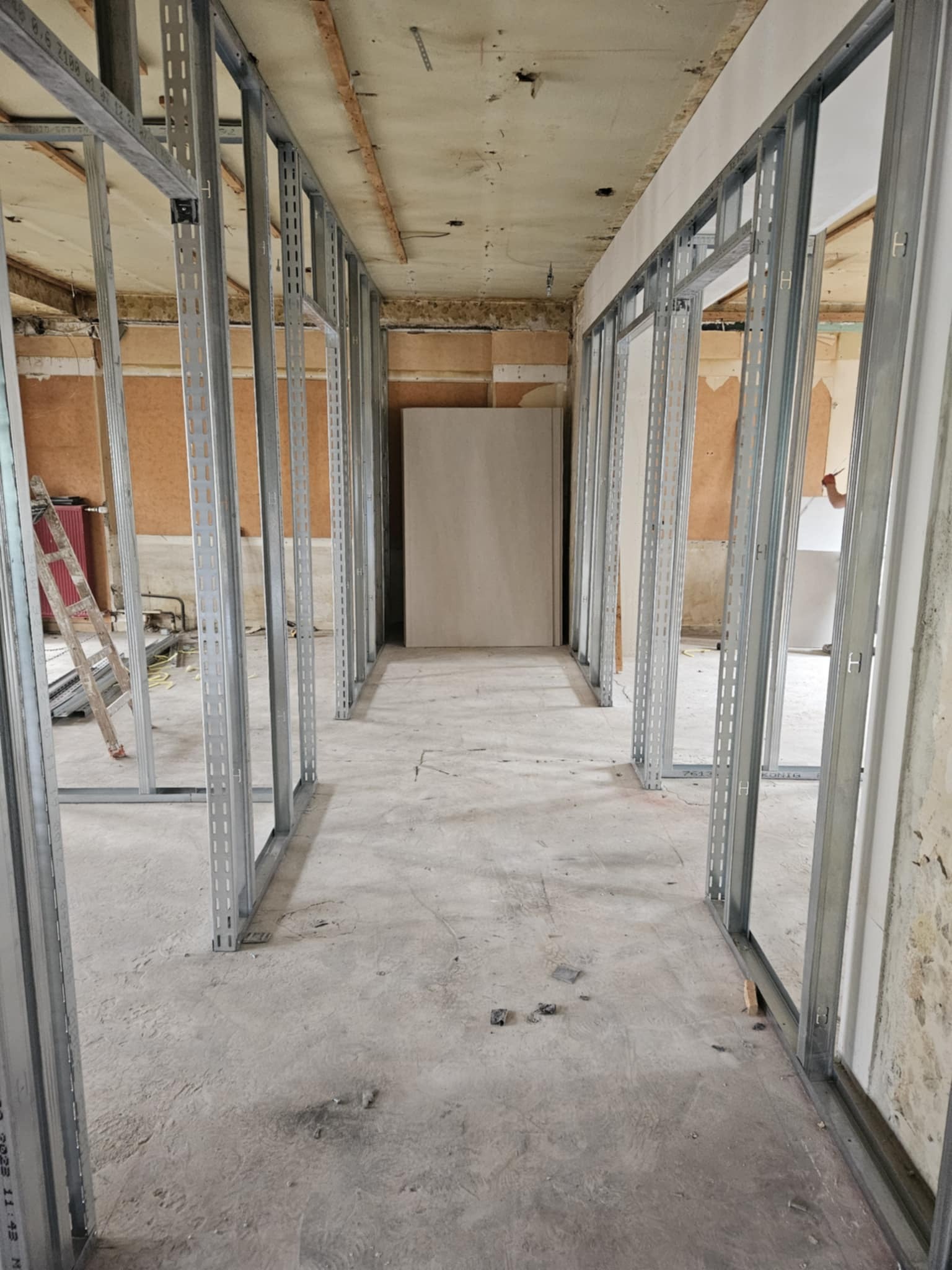 Korytarz w trakcie montażu ścian działowych z metalowych profili. Widoczne płyty karton-gips, drabina i surowy stan wykończenia wnętrza.