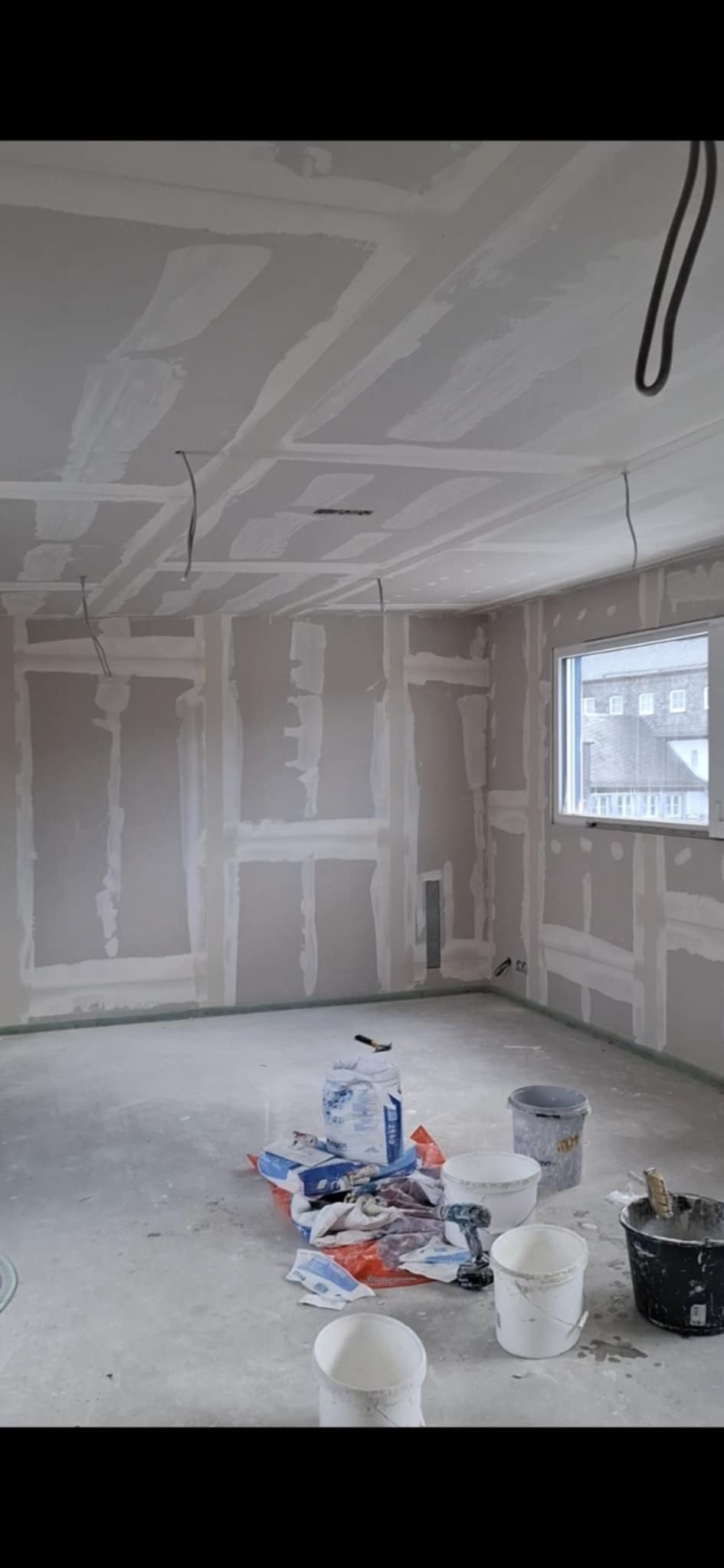 Pokój w trakcie prac wykończeniowych: ściany i sufit po szpachlowaniu, widoczne kable, worki z materiałami i narzędzia malarskie na podłodze. Okno z widokiem na budynek.