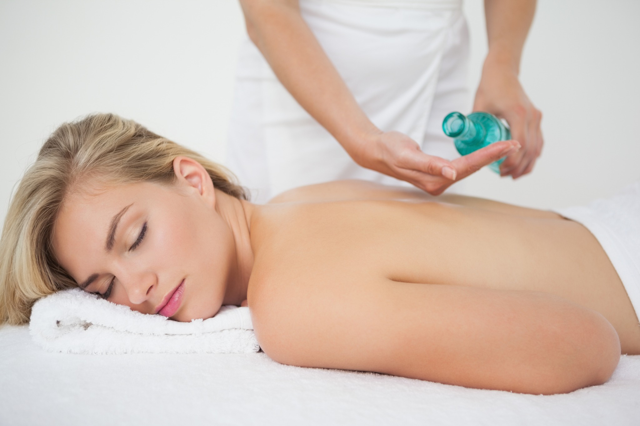 Kobieta relaksuje się podczas masażu pleców z użyciem olejku w gabinecie, w tle dłonie masażysty z butelką. Jasne wnętrze, miękkie światło.