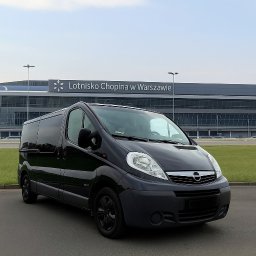 Szymon Walczak - Czarny bus Opel Vivaro na tle Lotniska Chopina w Warszawie, gotowy do przewozu osób. Pojazd z przyciemnianymi szybami i czarnymi felgami.