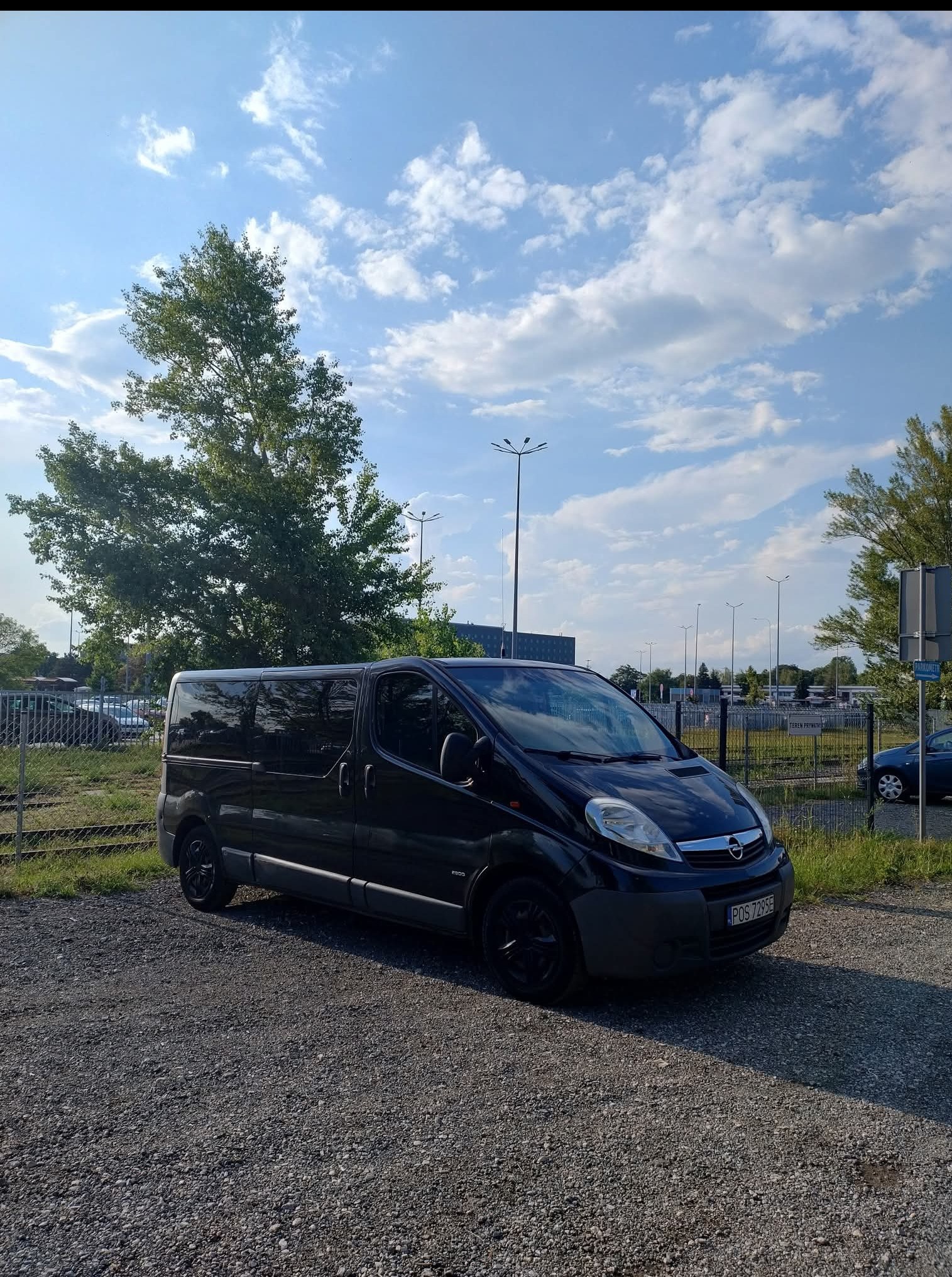 Czarny bus Opel Vivaro na żwirowym parkingu, wysoki kadr z widokiem na niebo i drzewa. Auto zaparkowane obok ogrodzenia, tablica rejestracyjna widoczna.