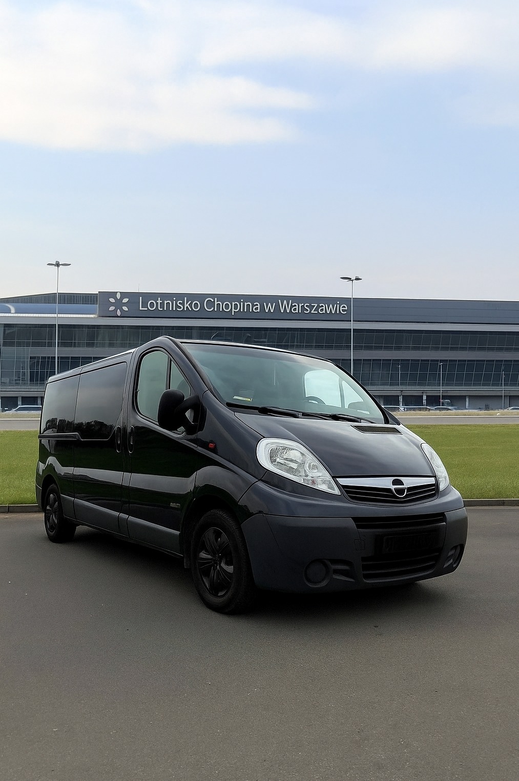 Czarny bus Opel Vivaro na tle Lotniska Chopina w Warszawie, gotowy do przewozu osób. Pojazd z przyciemnianymi szybami i czarnymi felgami.