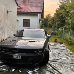 Czarny Dodge Challenger myty pianą na zewnątrz, na zielonej plandece, z myjką ciśnieniową Yato w tle, w otoczeniu budynków i zieleni, o zmierzchu.