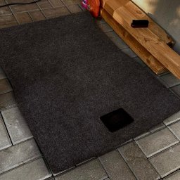 Mobilny Urban Shine Detailing - Ciemnoszary dywanik samochodowy z czarnym otworem, leżący na szarym bruku. W tle chemia i szczotka do czyszczenia.