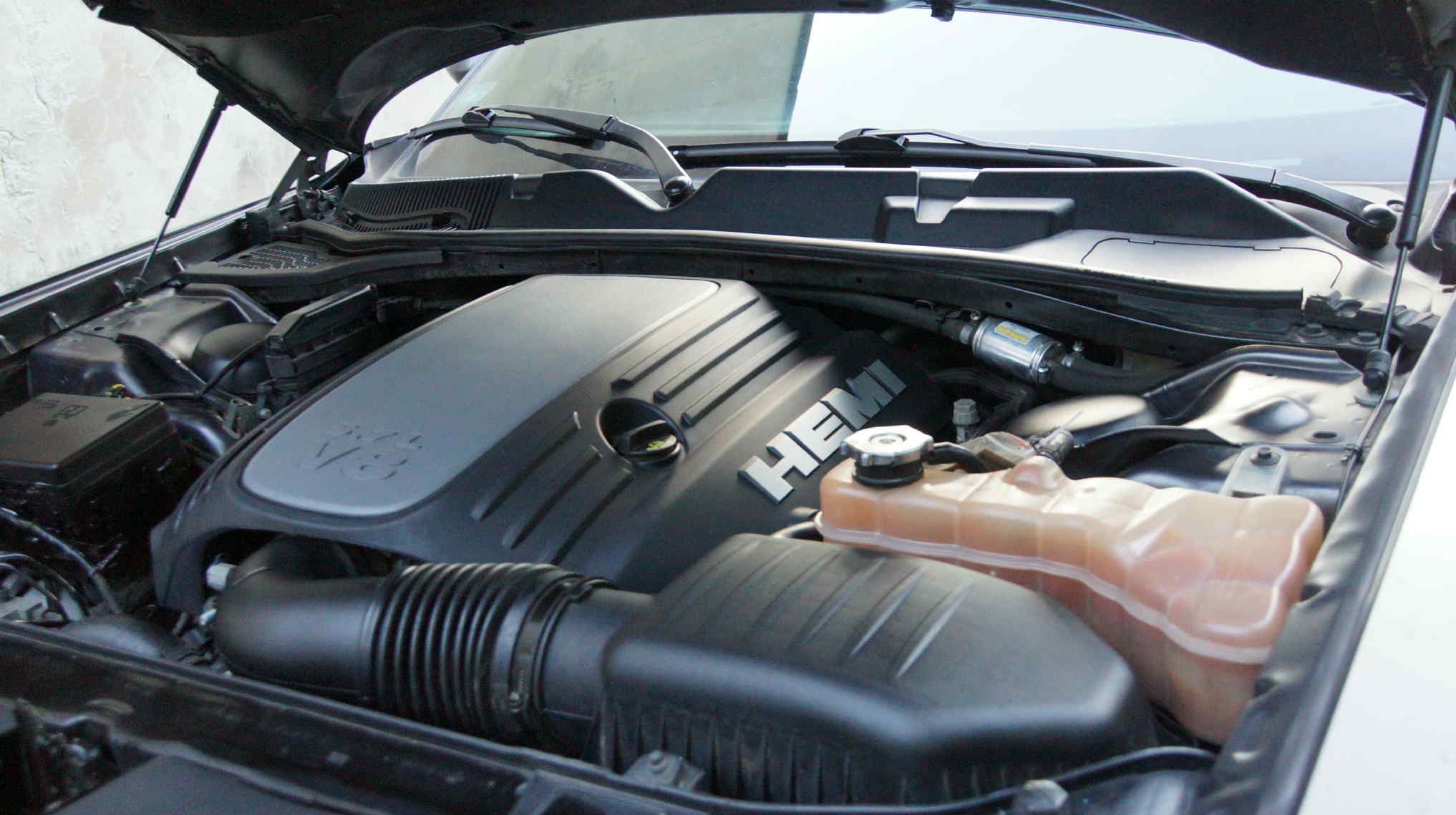 Otwarta maska czarnego samochodu z widocznym silnikiem V8 Hemi, zbiornikiem płynu chłodniczego i filtrem powietrza. Widoczne elementy instalacji elektrycznej i węże.