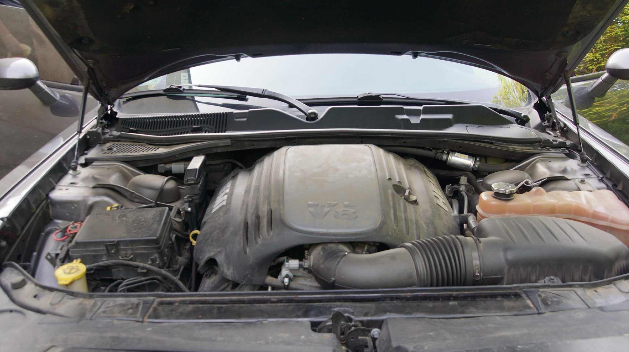 Otwarta maska samochodu, widoczny silnik V8. Elementy silnika lekko zabrudzone, w tle widoczne szyba i wycieraczki. Ujęcie z bliska na komorę silnika.