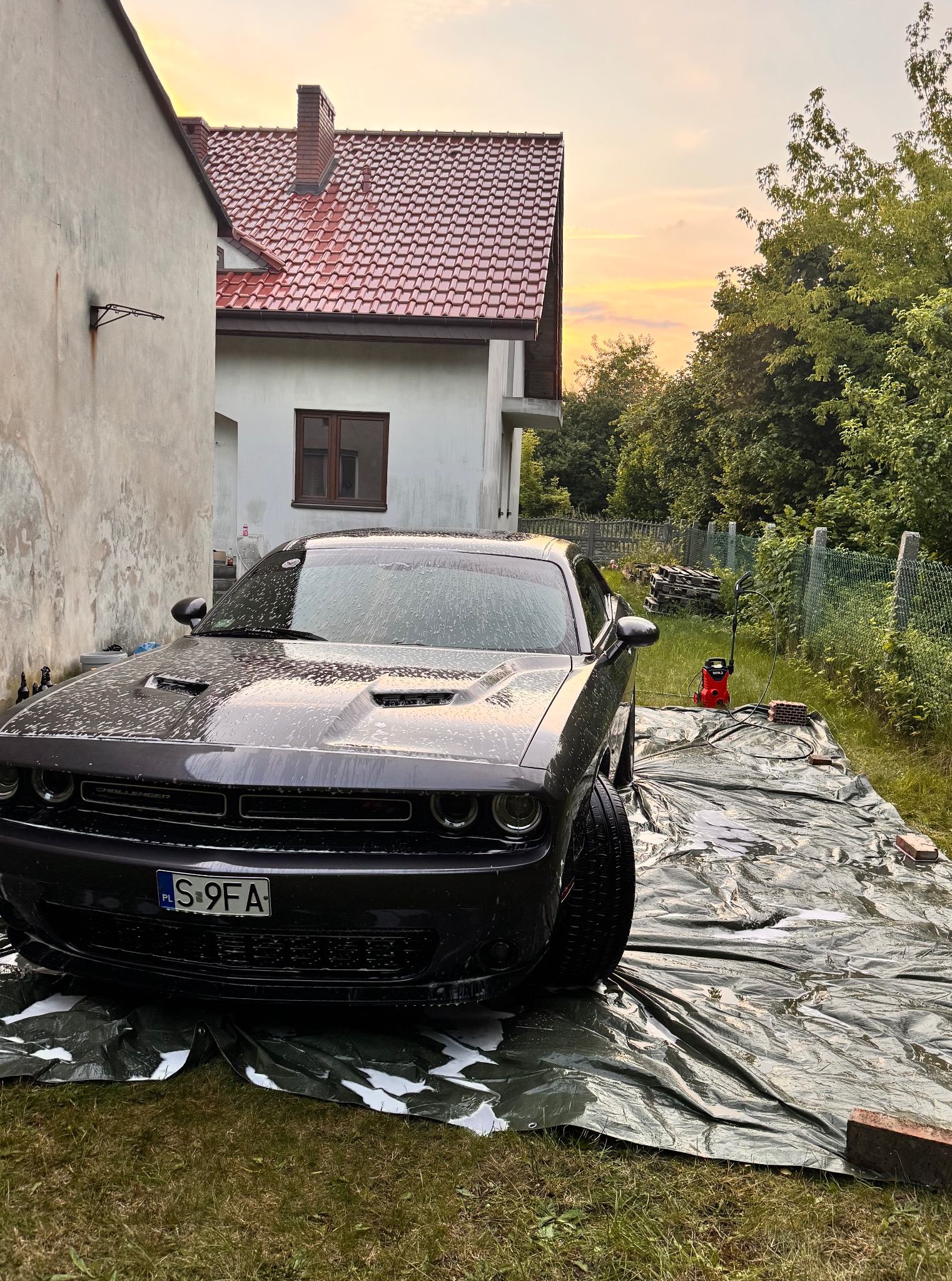 Czarny Dodge Challenger myty pianą na zewnątrz, na zielonej plandece, z myjką ciśnieniową Yato w tle, w otoczeniu budynków i zieleni, o zmierzchu.