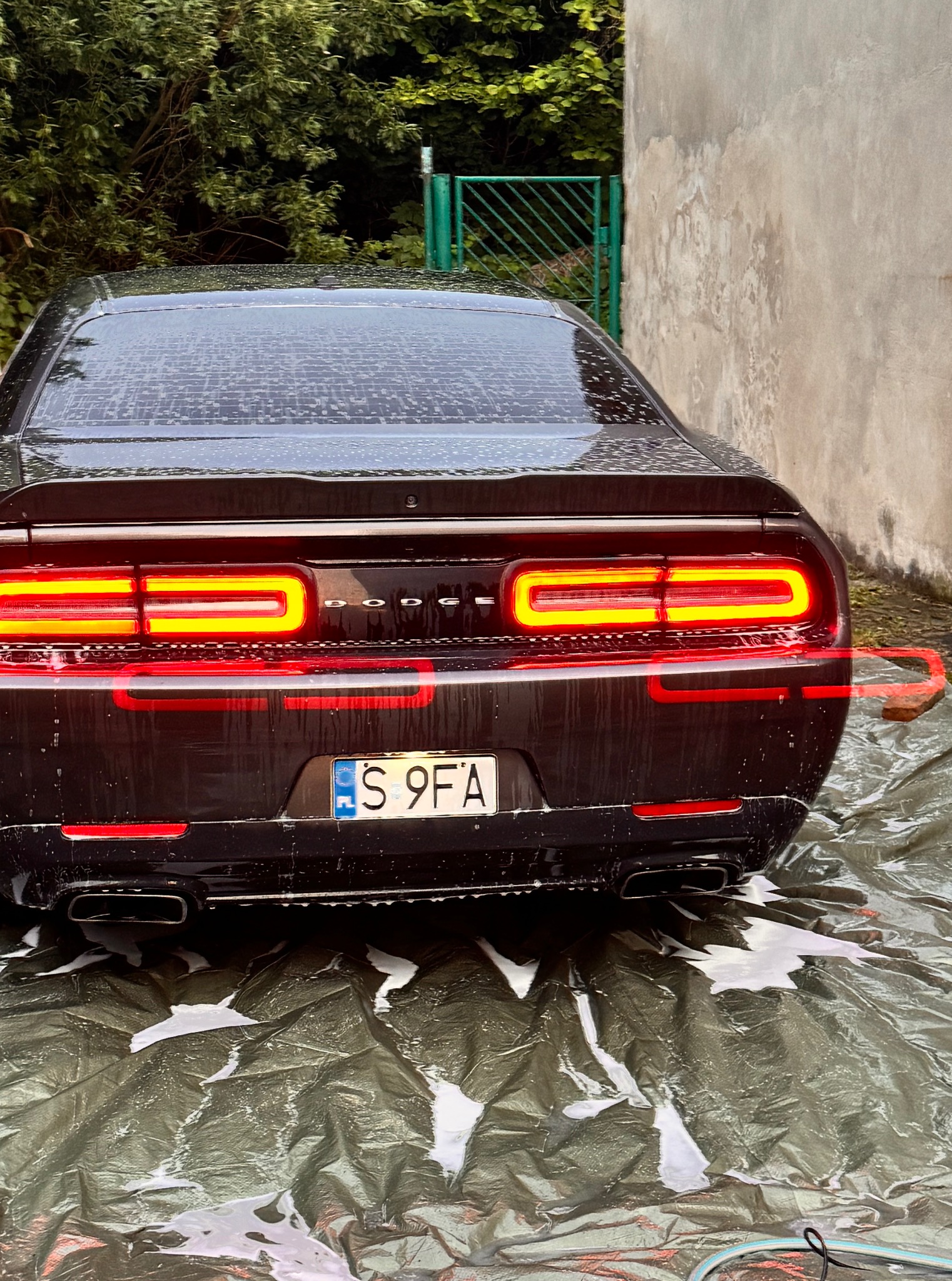 Czarny Dodge Challenger po myciu, widok z tyłu, mokry lakier odbija światło. Widoczne tylne światła, tablica rejestracyjna i resztki piany na plandece.