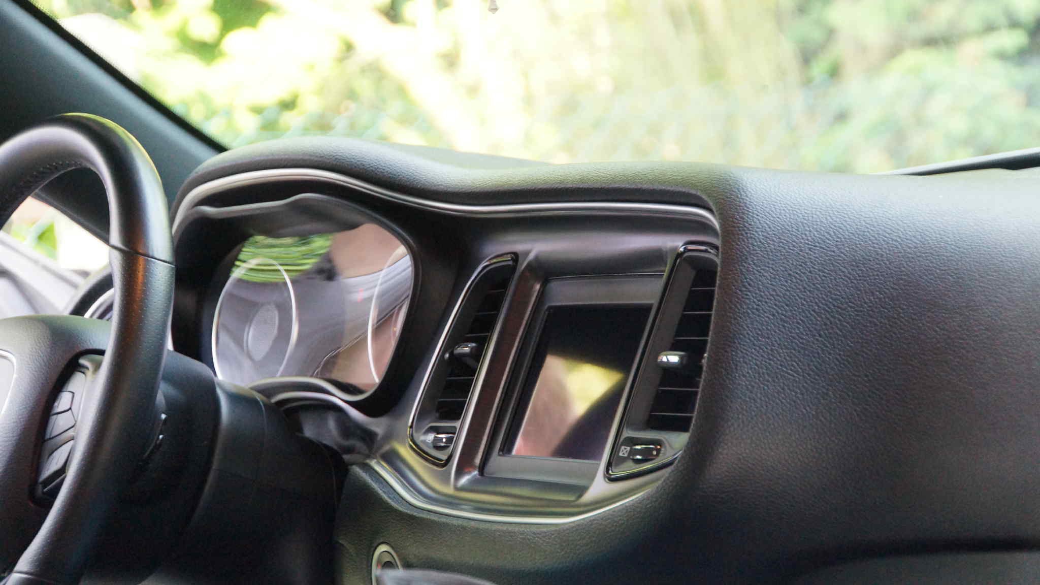 Czarna deska rozdzielcza auta z ekranem dotykowym i srebrnymi akcentami, widoczna kierownica. Tło rozmyte, zielone. Detal wnętrza pojazdu.