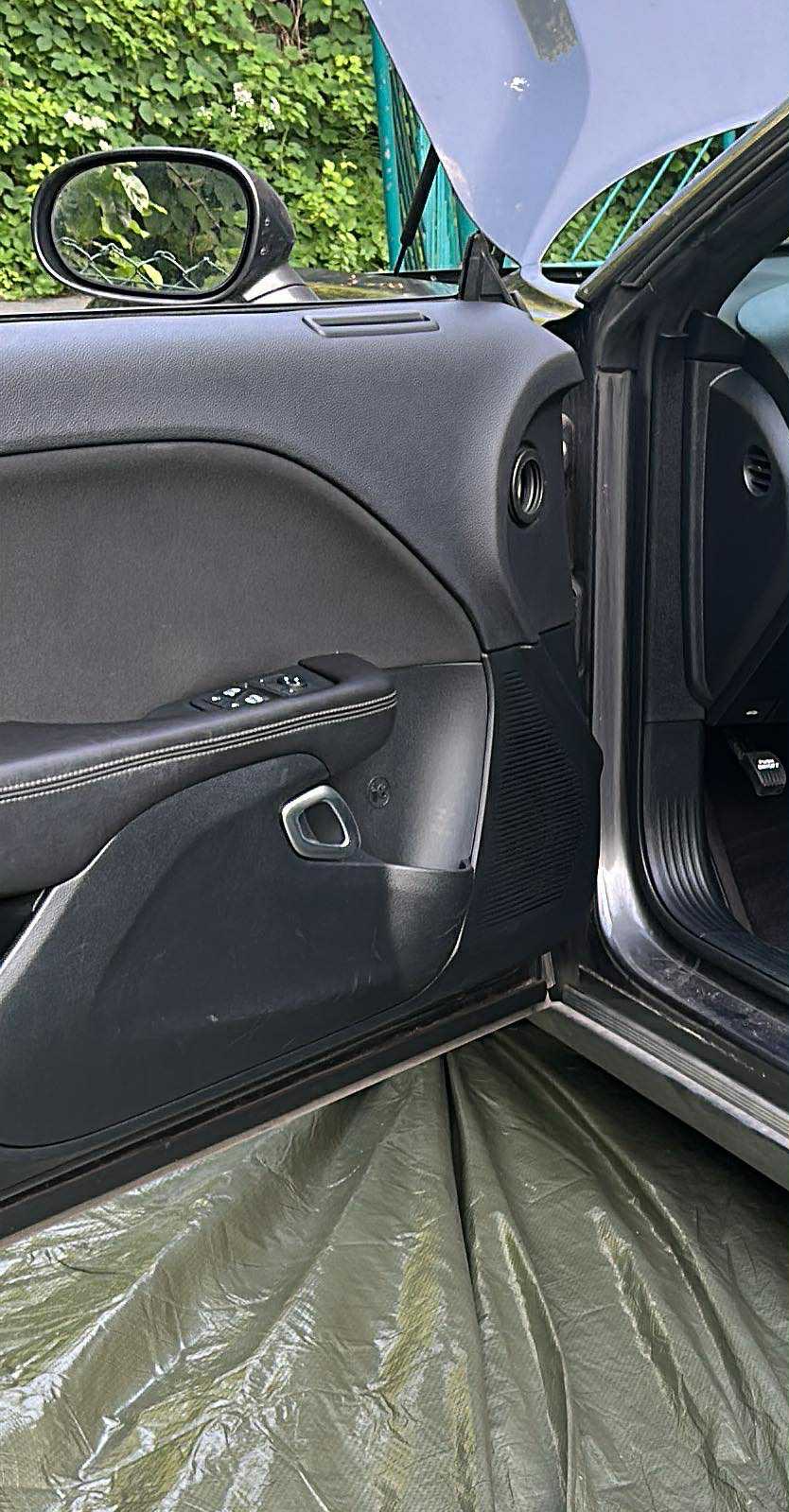 Wnętrze samochodu z otwartymi drzwiami, widoczny panel drzwiowy z elementami sterującymi szybami, lusterko boczne i fragment karoserii, zabezpieczone folią ochronną.