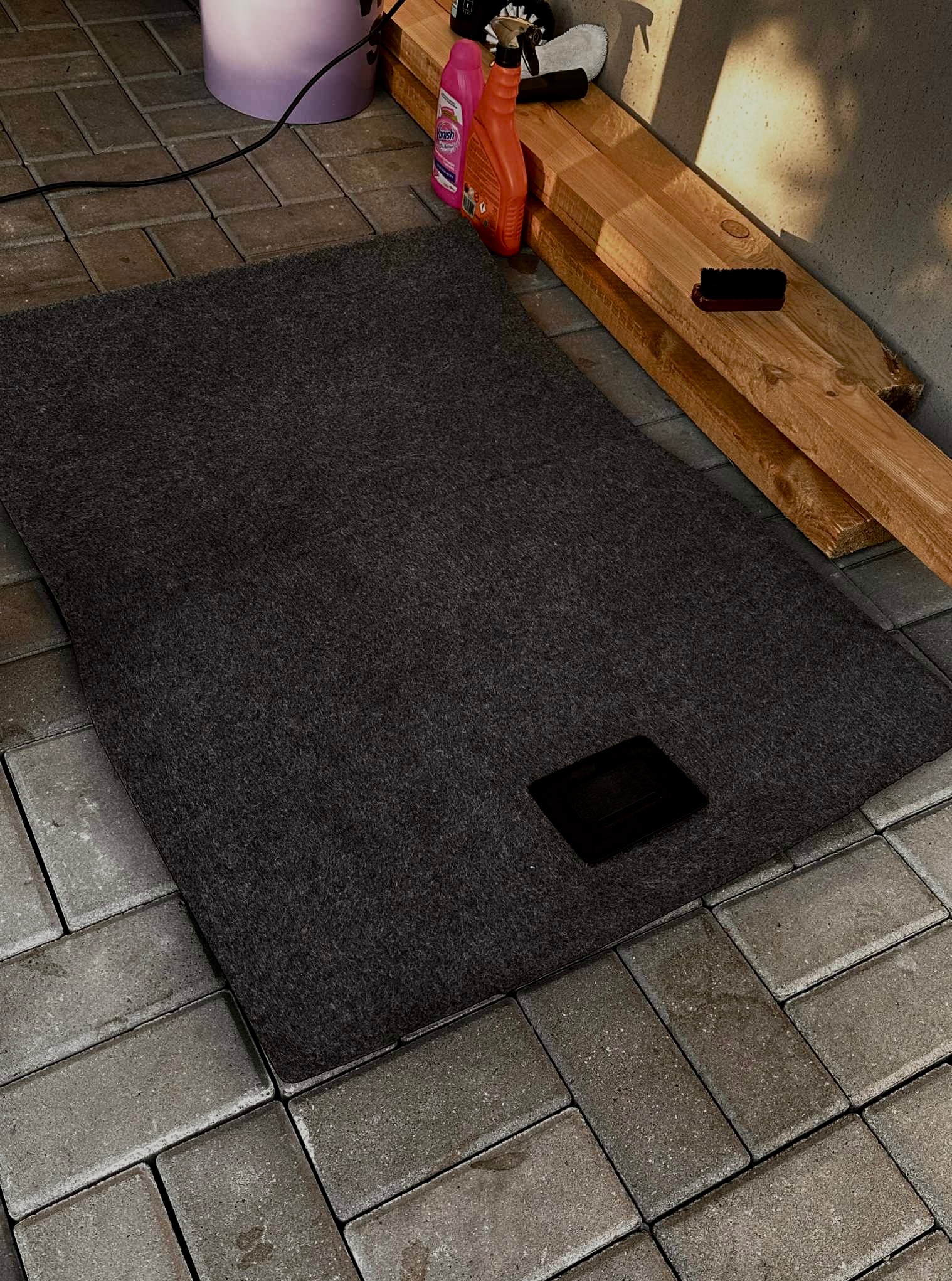 Ciemnoszary dywanik samochodowy z czarnym otworem, leżący na szarym bruku. W tle chemia i szczotka do czyszczenia.