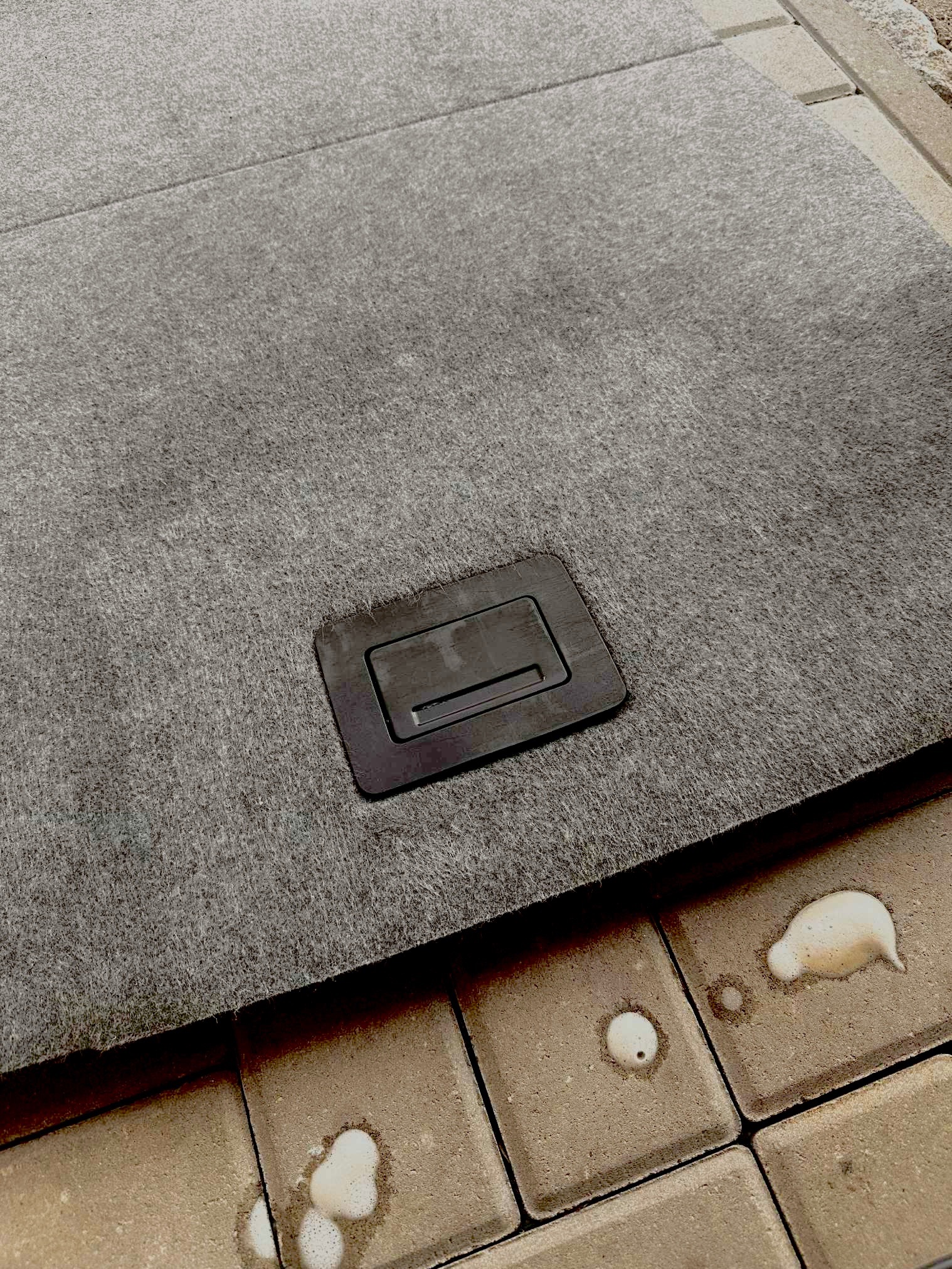 Szara, tekstylna wykładzina bagażnika samochodowego z czarnym uchwytem, widoczna na tle kostki brukowej z plamami. Detal wnętrza pojazdu.