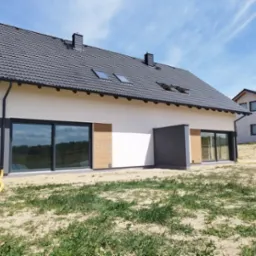 Wykończony dom jednorodzinny z jasną elewacją, grafitowym dachem i dużymi oknami przesuwnymi. Widoczny fragment ogrodu i sąsiedni dom w tle.