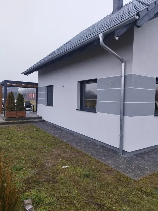 Ściana domu z szarymi pasami, rynną i czarnymi ramami okiennymi. Widoczny fragment dachu i trawnika. Minimalistyczny design elewacji w stonowanych kolorach.