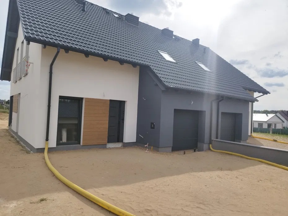 Dwukondygnacyjny dom z szarą dachówką, białą i szarą elewacją. Widoczne dwa garaże i drewniana okładzina. Żółty wąż leży na piaszczystym podłożu.