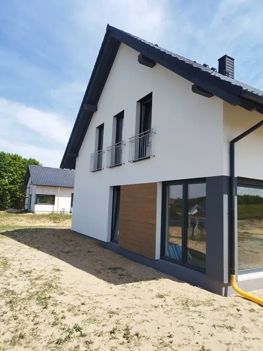 Nowoczesny dom jednorodzinny z białą elewacją, czarnym dachem i drewnianymi akcentami. Widoczne okna z balustradami i fragment ogrodu.