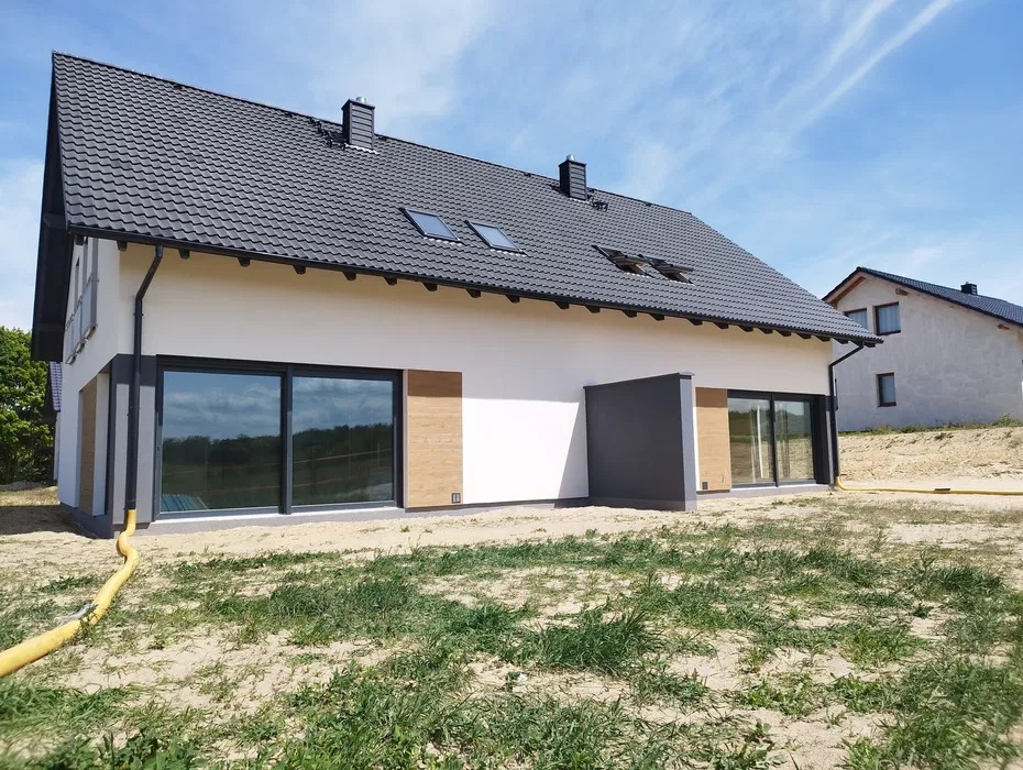 Wykończony dom jednorodzinny z jasną elewacją, grafitowym dachem i dużymi oknami przesuwnymi. Widoczny fragment ogrodu i sąsiedni dom w tle.