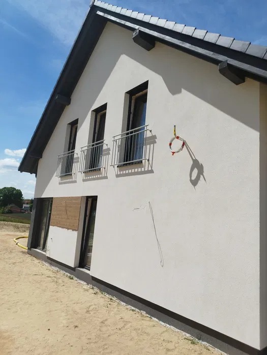 Jasna elewacja domu jednorodzinnego z trzema balkonami na piętrze. Widoczne fragmenty ocieplenia i instalacji. Świeżo otynkowana ściana w słoneczny dzień.