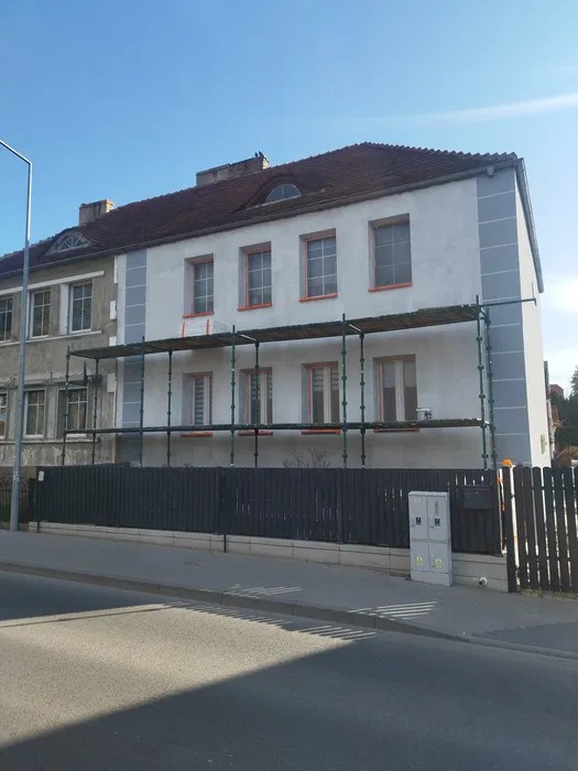 Elewacja budynku w trakcie renowacji, widoczne rusztowania i częściowo odnowiona fasada. Kontrast między starą a nową częścią ściany.